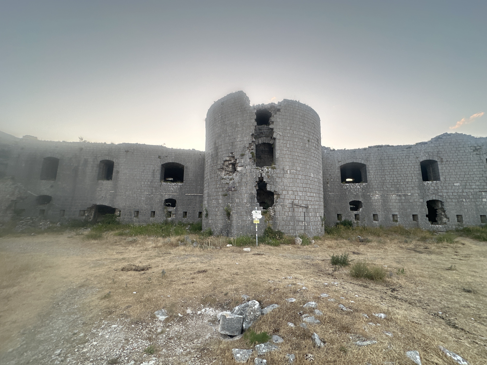 The image shows the ruins of a stone fortress in Brajici, Montenegro. The fortress is made of large, gray stones and has a central, cylindrical tower that is partially collapsed. There are several arched openings and smaller square windows along the fortress walls. The surrounding area is covered in dry, yellow grass and scattered rocks. The sky is overcast and hazy, suggesting it might be early morning or late evening. A signpost with directional arrows is visible in front of the central tower. The overall scene conveys a sense of abandonment and decay.