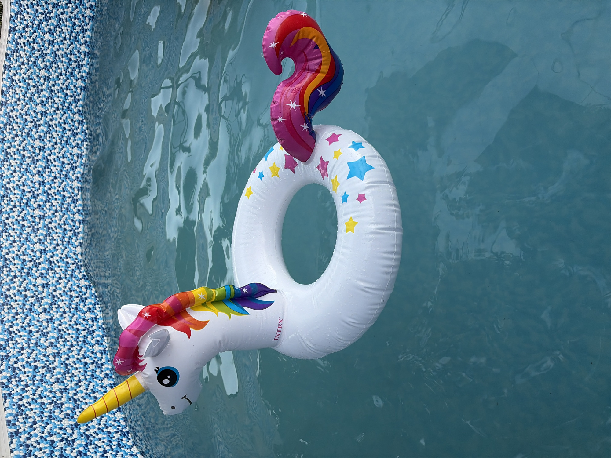 An inflated, white, unicorn-shaped pool float with a rainbow mane and tail floats in a swimming pool. The float has a yellow horn, blue eyes, and a pink mane with rainbow accents. Stars in various colors—pink, yellow, blue, and light blue—are scattered across the body of the float. The pool is filled with clear, blue water, and the tiled edge of the pool, with a pattern of small blue and white tiles, is visible at the top of the frame. The scene appears to be outdoors, possibly on a sunny day, judging by the bright lighting and the water's surface. There are no people present in the image. The location is described as Oxentea, Moldova.