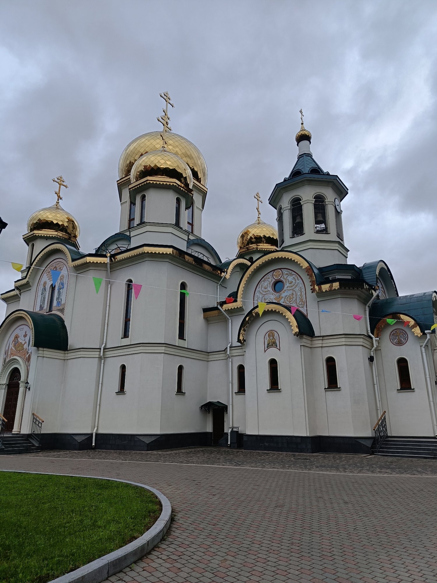 The image depicts the Church of the Holy Apostles Peter and Paul in Saint Petersburg, Russia. The architectural style is Orthodox. The church is predominantly white with golden domes topped with crosses. Decorative mosaics adorn parts of the exterior. Strings of colorful pennants are strung along the building. The sky is overcast and gray. A curved walkway made of patterned bricks leads to the church. A patch of green lawn borders the walkway.