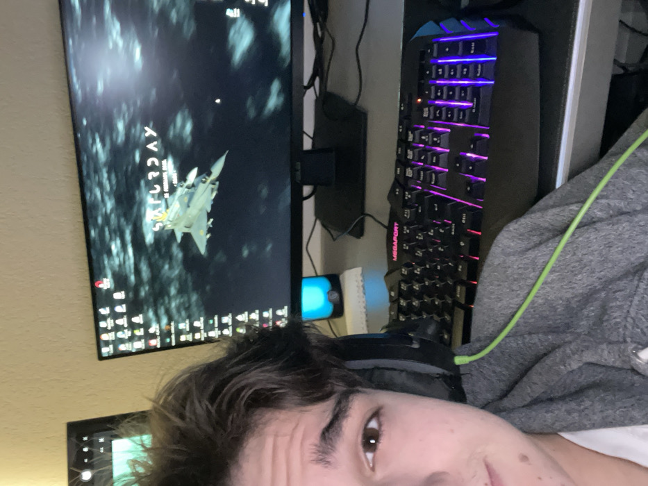 A young person's face is captured in a close-up, looking towards the camera from the lower right of the frame. They have brown eyes and dark, wavy hair, and are wearing a grey hooded sweatshirt with a bright green cord, likely from headphones, draped over it. Black over-ear headphones are visible on their head.

In the background, a computer monitor is prominently displayed, showing a dark desktop wallpaper. The wallpaper features a fighter jet, appearing to be a Saab JAS 39 Gripen, flying through a cloudy, dark sky. Text visible on the screen includes "SATURDAY" and "at 11:32 PM," which is part of the wallpaper design, and "4:11" in the top-left corner, likely a clock widget. Numerous application icons are arranged along the bottom of the monitor.

To the right of the monitor is a gaming keyboard, illuminated with vibrant RGB backlighting in purple and blue tones. The brand "MEGAPORT" is visible on the keyboard. A device emitting a bright blue light, possibly a smartphone or small speaker, is situated between the monitor and the keyboard. Multiple cables and wires are visible behind the computer setup, suggesting a workstation. The background wall is a plain, light color.