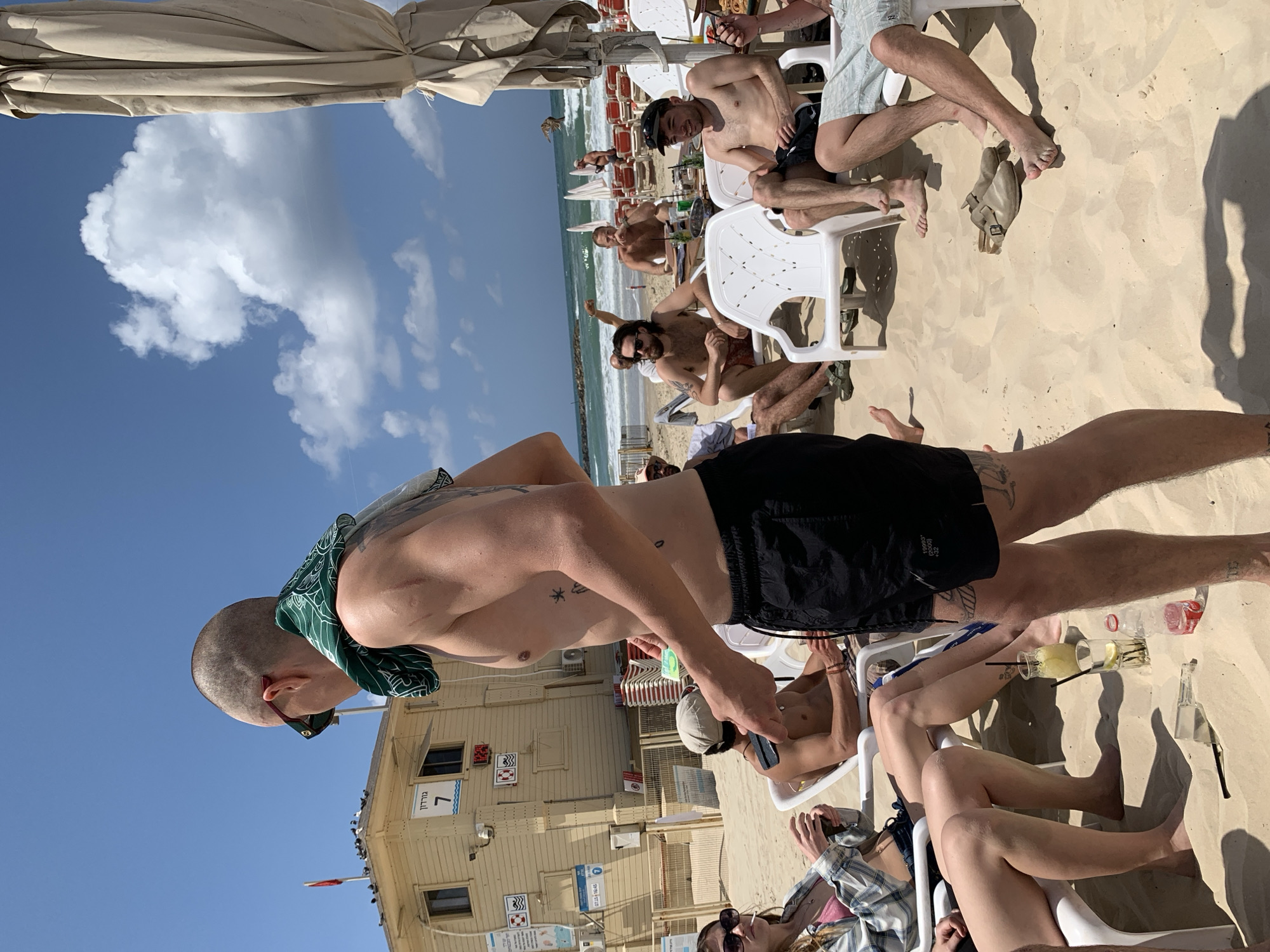 This media file captures a vibrant, sunny beach scene in Tel-Aviv, Israel. The setting is a light-sanded beach with the Mediterranean Sea stretching into the distance under a bright blue sky dotted with white cumulus clouds.
In the foreground, a man with a shaved head, wearing sunglasses and a green patterned bandana, stands shirtless with black swim shorts. He is seen from the back, revealing tattoos on his arms and upper back. His posture suggests he is either observing the scene or interacting with others.
Behind him, several individuals, mostly men, are relaxing on white plastic chairs on the sand. Some are shirtless, while others wear casual beach attire. They appear to be socializing and enjoying drinks; a glass of lemonade and other bottles are visible. One man in a black cap looks directly towards the camera with a smile. Another person is partially visible in the lower right, wearing a plaid shirt and sunglasses.
In the background, rows of collapsed beach umbrellas and more chairs line the shore. A light-colored, wooden-paneled beach building, likely a lifeguard station or facility, stands prominently. On its facade, a large red number "7" is clearly visible, along with Hebrew text above it that reads "גורדון" (Gordon), indicating "Gordon Beach". Various international beach safety symbols are also present on the building. The overall atmosphere is one of relaxation and leisure on a bright, active beach day.