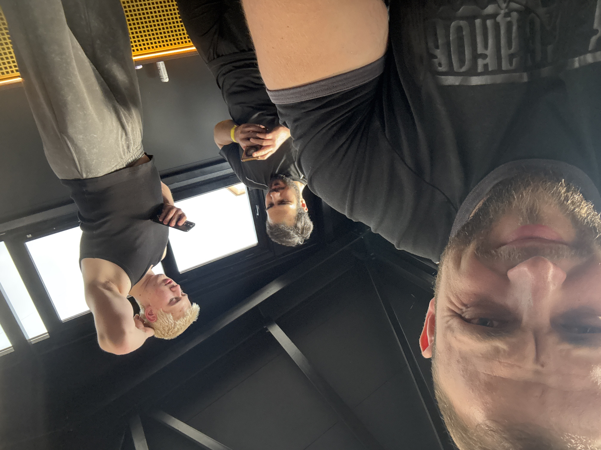 The image, originally rotated 90 degrees counter-clockwise, presents a low-angle perspective looking up at three men in an indoor setting.
In the foreground, a man with a beard and light-colored hair is partially visible, his face prominent as he looks downwards with an intense or squinting expression, showing wrinkles around his eyes. He wears a dark t-shirt, on which stylized text "ROHUR" is visible, likely a mirrored or reversed form of "POWER".
Further back and to the left, a second man with dark hair and a beard is seen wearing a black t-shirt and black pants. He holds a phone in his hands and also appears to be looking down. A yellow wristband adorns his left wrist.
To the far left, a third man with short, bleached blonde hair is dressed in a black sleeveless tank top and light grey athletic pants. He holds a phone and looks away from the camera, revealing a muscular left arm.
The background features a dark ceiling with visible black beams or support structures, suggesting an industrial or modern architectural style. Bright, rectangular windows or skylights are visible overhead, providing natural light to the space. A yellow, textured or gridded surface is also present in the upper background, possibly a decorative element or part of gym equipment.
The men appear to be casually standing or taking a break, possibly checking their phones. The unique perspective makes them seem to be looking down at the viewer. The overall atmosphere suggests a daytime setting, likely in a gym or a contemporary communal area.