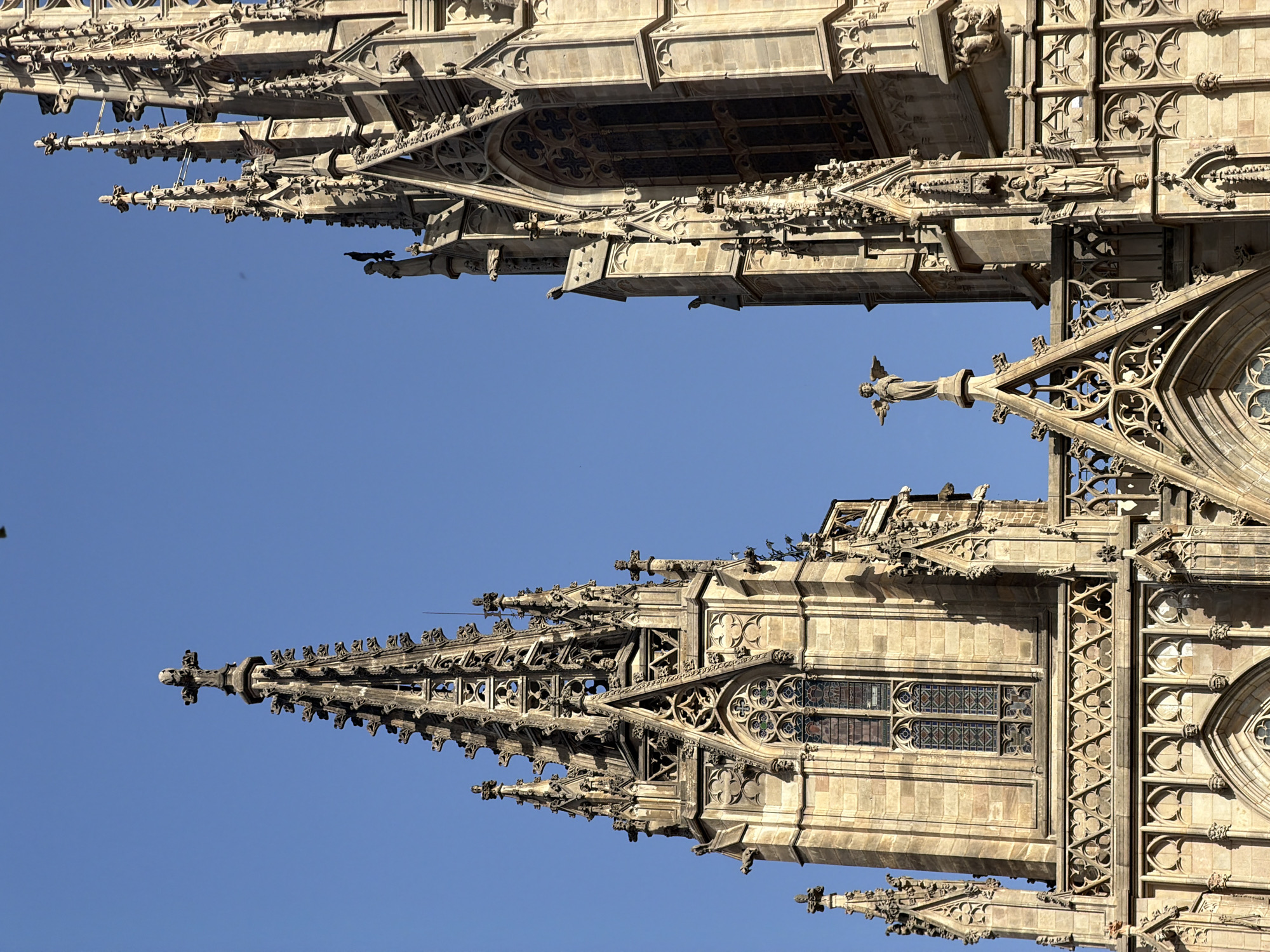 The image shows the intricate gothic architecture of the Barcelona Cathedral in Barcelona, Spain. The view is a close-up of two of the cathedral's spires against a clear blue sky. The spires are made of stone and are adorned with numerous decorative elements, including pinnacles, gargoyles, and ornate carvings. Stained glass windows are visible within the spires. A small statue of an angel with outstretched wings is perched on a finial between the two spires. Birds are present, perched on ledges and in flight, contributing to the realistic depiction of the scene. The stone appears weathered, suggesting age and exposure to the elements. The sunlight casts shadows that highlight the detailed stonework and three-dimensional quality of the cathedral.