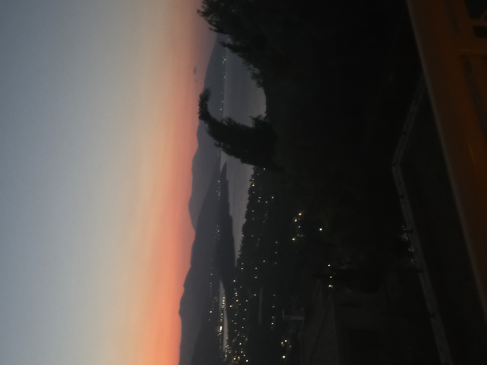 The image depicts a twilight scene from a vantage point overlooking a coastal area in the Municipal Unit of Asini, Greece. The sky transitions from a soft orange and pink hue near the horizon to a pale blue higher up. In the mid-ground, rolling hills or mountains are silhouetted against the sky. Nestled along the coast, a town or village is visible, illuminated by numerous scattered lights that twinkle in the fading daylight. A body of water, likely the sea, reflects the dim light and stretches towards the distant hills. In the foreground, dark silhouettes of trees and possibly some structures are visible, suggesting a natural or built-up environment from which the photograph was taken. The overall atmosphere is serene and peaceful, characteristic of dusk.