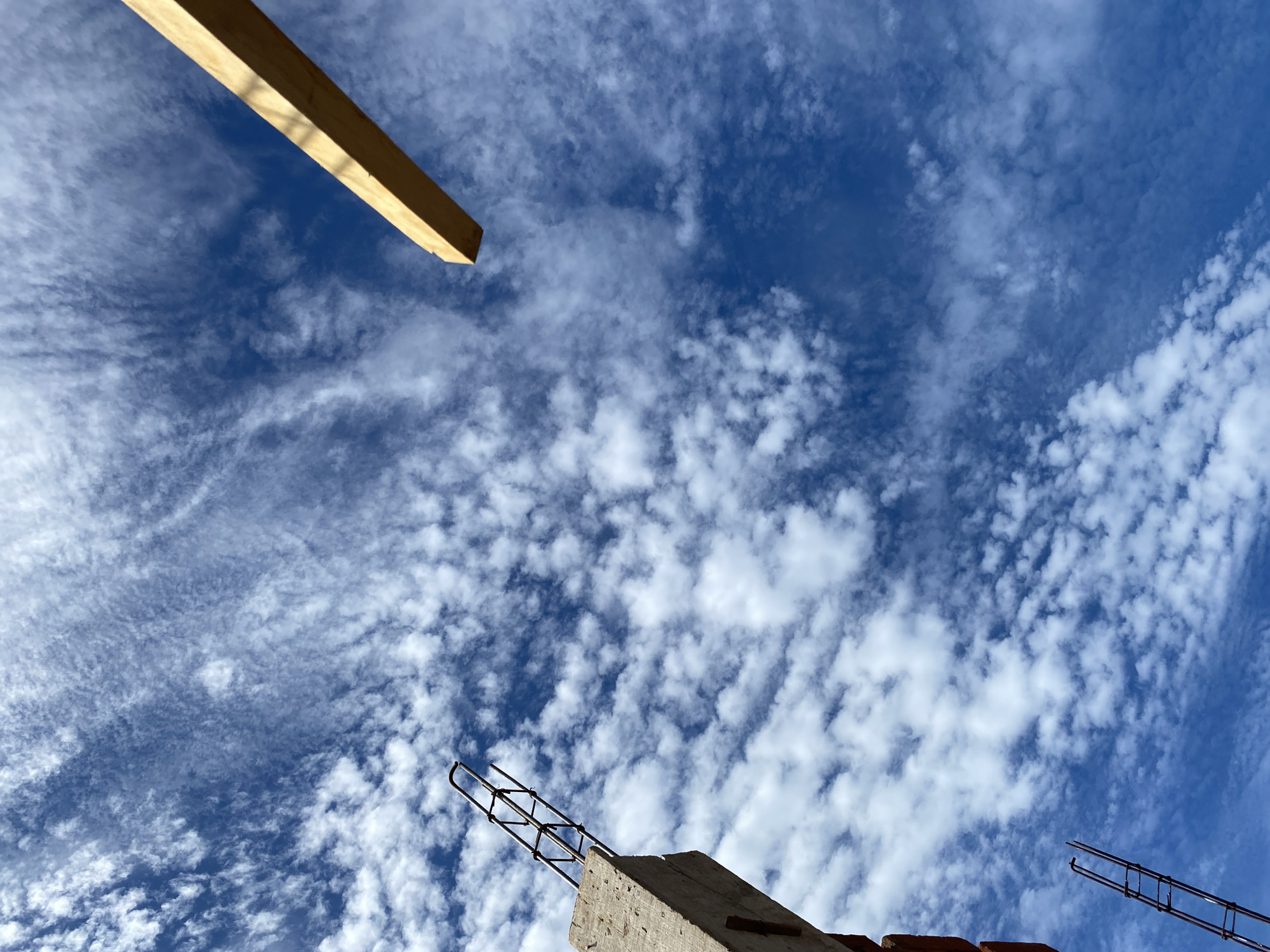 An outdoor, low-angle shot captures a construction site against a bright blue sky filled with wispy white clouds. In the lower left, a concrete pillar with rebar protruding from the top stands vertically, partially obscured by brickwork. Further down and to the left, another section of rebar scaffolding is visible. In the upper right quadrant, a light-colored wooden beam extends diagonally from the top right corner into the frame. The scene is devoid of people, indicating an inactive construction phase. The weather appears clear and sunny. The image does not provide enough information to determine the specific location within Banda Aceh, Indonesia.