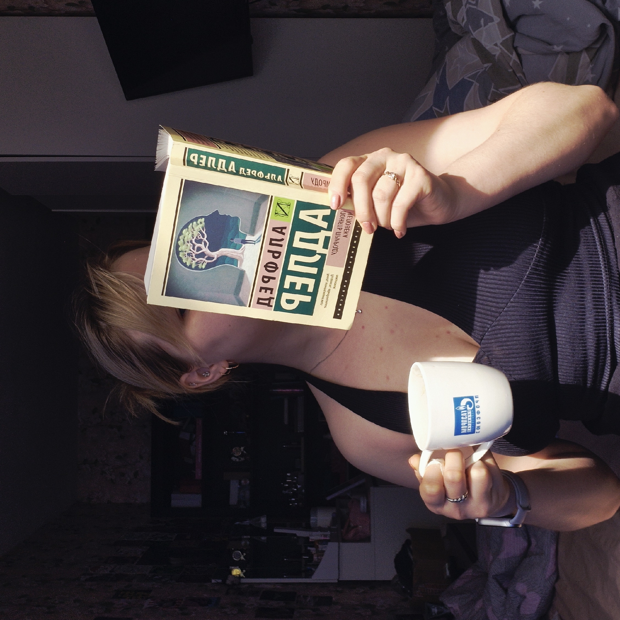 The image shows a person holding a book and a mug. The book is titled "ДЭФФЕР" (DEFFER) by Дэрфрора (Derfrofa) in Russian, and the book's cover features an image of a tree inside a silhouette of a head. The mug has a logo and says "МОЧПЕАТ ГАЗПРОФСОЮЗ" which, when translated from Russian, likely refers to an organization, maybe related to "Gazprom" (a large energy company). The person is wearing a black top and has an earring. There's a smart watch on their wrist and rings on their fingers. In the background, there's a room with a bed and other furniture. The environment suggests an indoor setting, possibly a home or apartment. Based on the logo and the Russian text, the location could be in Russia. However, based on the information provided there is no indication that this image relates to the city of Chaikovsky.