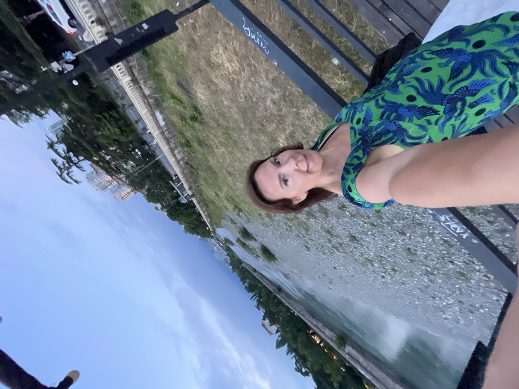 A woman is taking a selfie on a bridge over a river in Sochi, Russia. She is wearing a green dress with a blue bird pattern. The river is shallow and rocky, with a concrete embankment on the left side. In the background, there are trees, buildings, and a white car on a road. The sky is overcast.