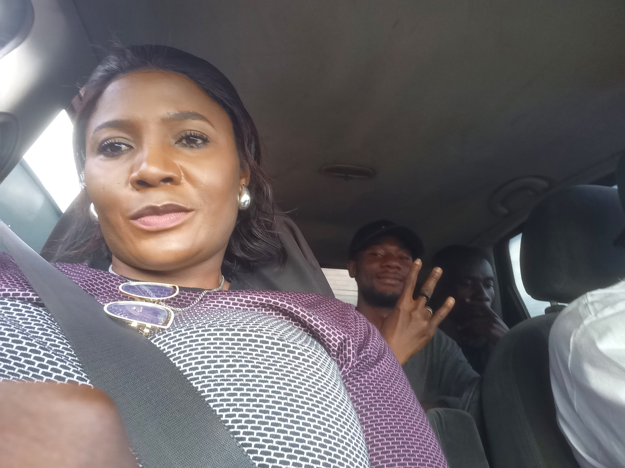 The image is a selfie taken from inside a vehicle, showing three individuals. A woman is in the foreground, seated in the driver's seat, wearing a purple and black patterned top and a necklace with two oval pendants. Her left arm is visible, and she is wearing a seatbelt. Behind her, to her right, a man wearing a black baseball cap is partially visible, making a peace sign with his right hand. Further back and to the right, another man is visible, with his head slightly lowered. The interior of the car appears to be a typical passenger vehicle, with a dark-colored headliner and visible seatbacks. The lighting suggests it is daytime, likely outdoors, as evidenced by the bright light entering the car from the left side. The background is blurry, indicating the vehicle is likely in motion. No specific city or location is identifiable from the image itself.