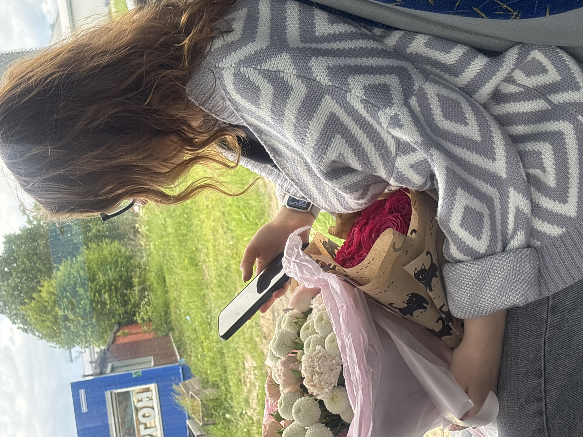 A person is seated in a bus, looking at a cell phone, while holding two bouquets of flowers. The person is wearing a grey and white sweater and has long brown hair. The bouquet on the left is white chrysanthemums wrapped in pink tissue paper. The bouquet on the right is red roses wrapped in brown paper. Through the bus window are green trees and grass, and part of a blue building with the letters "ГТОН" on it. The location is Unknown, Unknown.