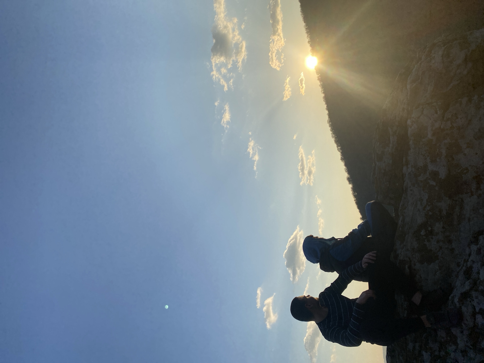 The image captures a picturesque outdoor scene, likely taken during either sunrise or sunset in the natural landscape of Goritsa, Bulgaria. Two individuals are prominently featured in silhouette, sitting on a rugged, rocky outcrop in the lower-right portion of the frame.
The person in the foreground wears a striped long-sleeved shirt and dark pants, while the second individual, positioned slightly behind and above, is dressed in a hooded jacket (appearing blue) and dark pants. Their features are obscured by the intense backlighting from the sun, but their relaxed postures suggest they are observing the expansive view.
The sky occupies a significant portion of the image, displaying a gradient from a deep blue on the left to a warm, golden hue towards the right where the sun is situated. Scattered, wispy white clouds are visible, catching the light and adding texture to the sky. The sun, a brilliant orb, is low on the horizon on the right side, partially concealed by a distant tree line or forested hill, casting radiant sunbeams across the scene. This 'golden hour' lighting creates strong contrasts and a serene, contemplative atmosphere. A continuous line of trees forms a dark silhouette along the distant horizon, indicating a dense forest or mountainous terrain. There is no visible text in the media file. The overall scene evokes a sense of peace and appreciation for nature's beauty.