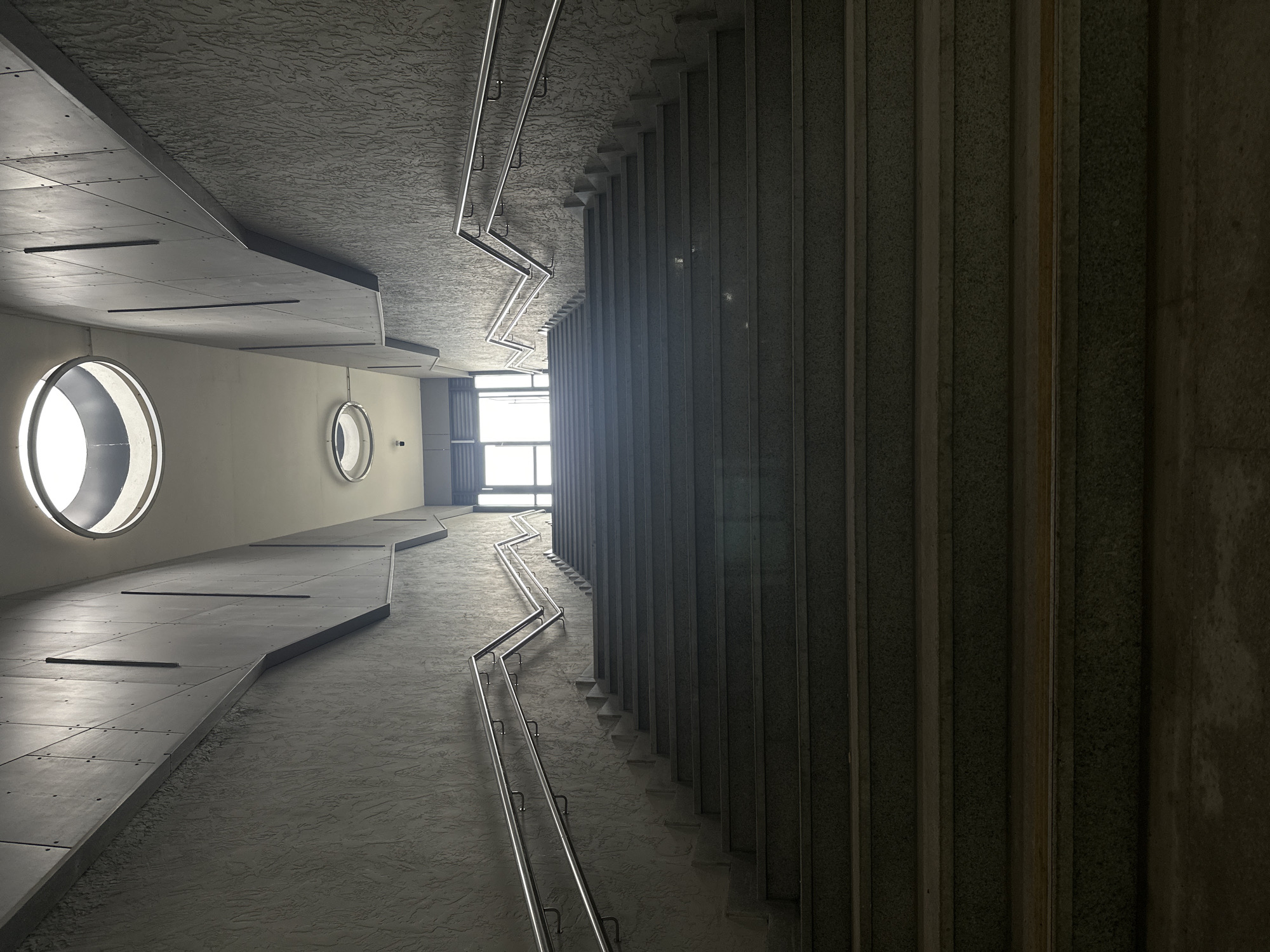 The image shows an interior view of an inclined walkway or ramp at the Kazan Family Center in Kazan, Russia. The space is characterized by modern architectural design. A wall to the left features large, circular windows. The walkway has a textured surface and is bordered by metallic paneling. A series of vertically-oriented, textured panels line the right side of the walkway. A stainless steel handrail runs along the center, following the incline. The walkway leads towards a bright, windowed area at the far end. No people are visible in the image.