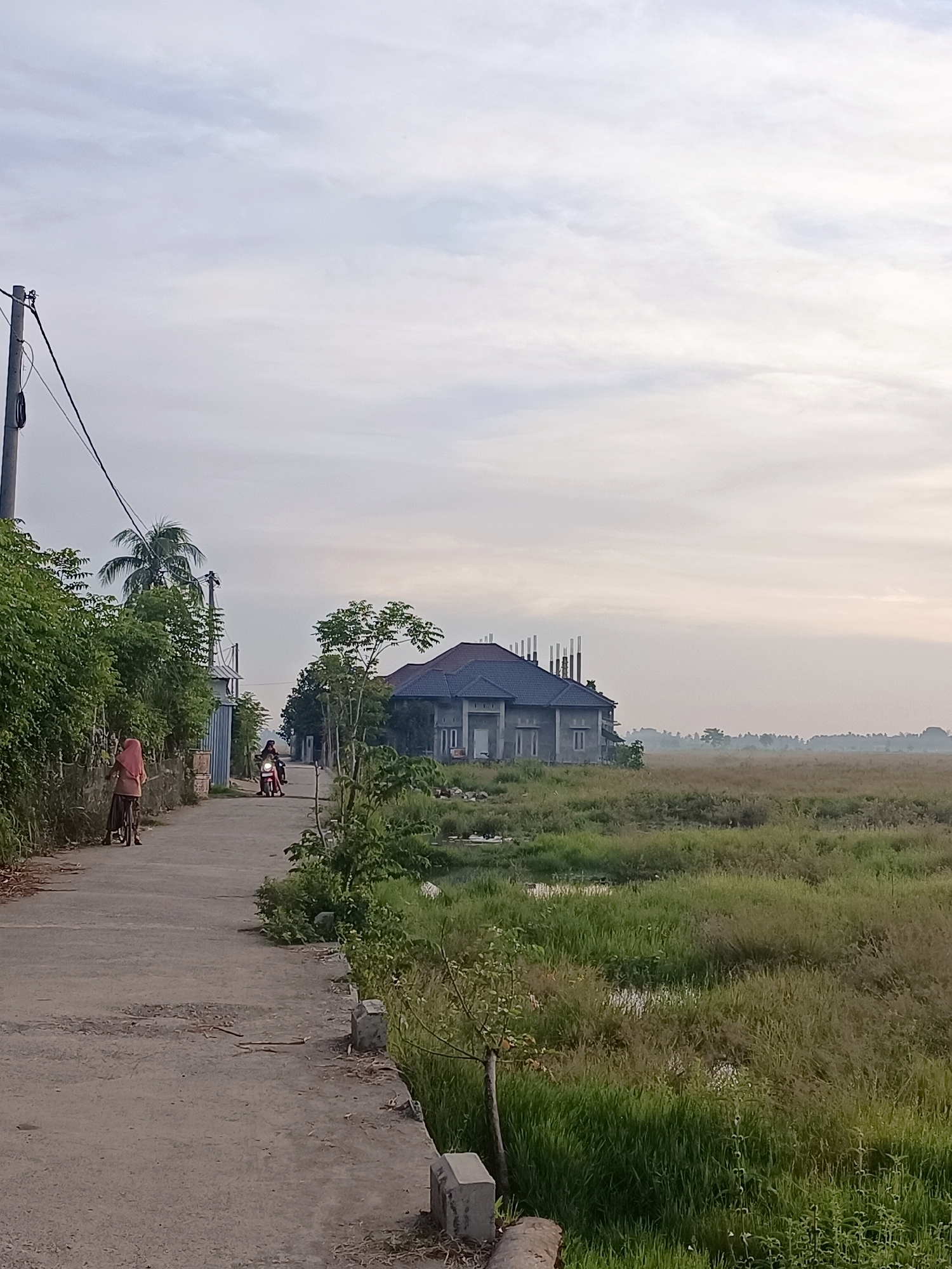 A concrete road with some cracks runs through a rural area in Meunasah Raya, Indonesia, under a pale, cloudy sky. On the left side of the road, dense green vegetation lines the path, with a utility pole and wires overhead. A person wearing a pink hijab and a brown skirt is walking a bicycle on the left side of the road. Further down the road, another person is riding a motorcycle. On the right side of the road, a large, unfinished house with a dark tiled roof and exposed concrete pillars is situated amidst a field of tall green grass. The field extends to the horizon, where a faint treeline is visible. The overall impression is one of a tranquil, developing rural landscape.