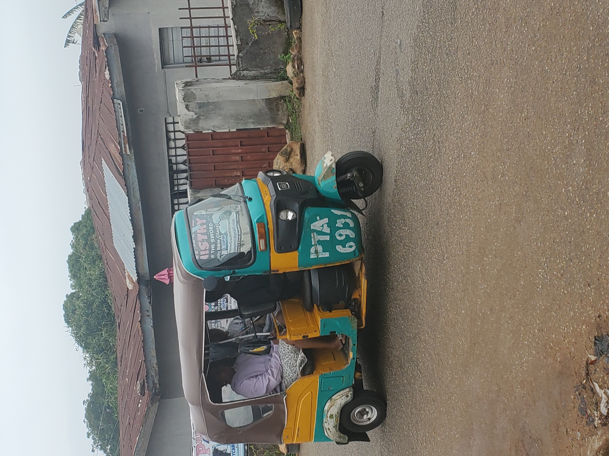 A stationary, three-wheeled motorized vehicle (a "keke"), parked on a paved road next to a building. The keke is painted in green, yellow and brown colors with license plate "PTA 6931". Passengers are seated within the vehicle. Advertisements are on the vehicle for a ministry "Throne of Graceintl Gospel" (location given as "Cows First Gaf. Jos", Nigeria). The area has a humid, overcast environment.