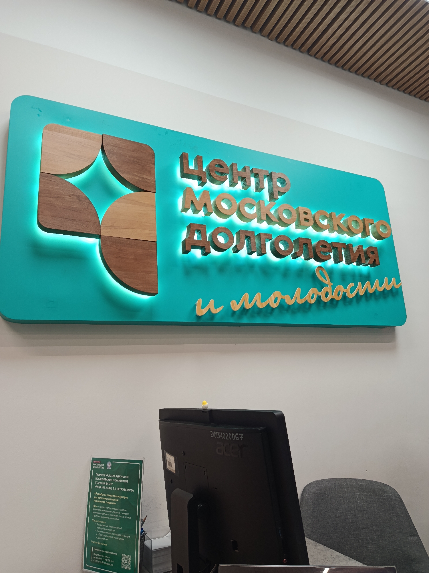 The image captures an indoor scene, likely a reception or information area within a facility in Moscow, Russia. The main subject is a large, brightly illuminated sign mounted on a light-colored wall.
The sign is teal-colored and features prominent 3D wooden letters that spell out "Центр Московского Долголетия и Молодости" (Center of Moscow Longevity and Youth). The letters are rendered in varying shades of light and dark wood and are backlit with a teal glow, creating a striking halo effect. To the left of the text, an abstract logo made of four wooden quarter-circles, also backlit, adds to the design. The ceiling above the sign has decorative wooden slats.
In the foreground, a black Acer computer monitor, bearing the serial number "210134020067" on its back, is visible on a desk. Part of a gray fabric chair can be seen behind the monitor. To the left of the monitor, a transparent acrylic stand displays a green-colored poster.
The poster, titled "МОСКОВСКОЕ ДОЛГОЛЕТИЕ" (Moscow Longevity), announces a scientific study: "ПРИМИТЕ УЧАСТИЕ В НАУЧНОМ ИССЛЕДОВАНИИ МЕХАНИЗМОВ СТАРЕНИЯ ФГБНУ «РНЦХ ИМ. АКАД. Б.В. ПЕТРОВСКОГО»" (Take part in a scientific study of aging mechanisms FGBNU "RSCM named after Acad. B.V. Petrovsky"). The study aims at "Разработка панели биомаркеров для комплексной оценки механизмов старения" (Development of a panel of biomarkers for comprehensive assessment of aging mechanisms), with the goal of "создать метод, который позволит оценивать особенности старения у каждого человека отдельно и подбирать персональные стратегии здорового долголетия" (creating a method to assess individual aging characteristics and select personalized healthy longevity strategies). Participants will receive "Расширенный биохимический и общий анализ крови," "Расчет биологического возраста," and a "Персональную карту здоровья и долголетия" (extended biochemical and general blood tests, biological age calculation, and a personal health and longevity map). It specifies "Участие бесплатное" (Participation is free) and notes "Имеются противопоказания!" (There are contraindications!). Information for registration is provided, indicating availability "с 9:00 до 16:00 в будние дни" (from 9:00 to 16:00 on weekdays).