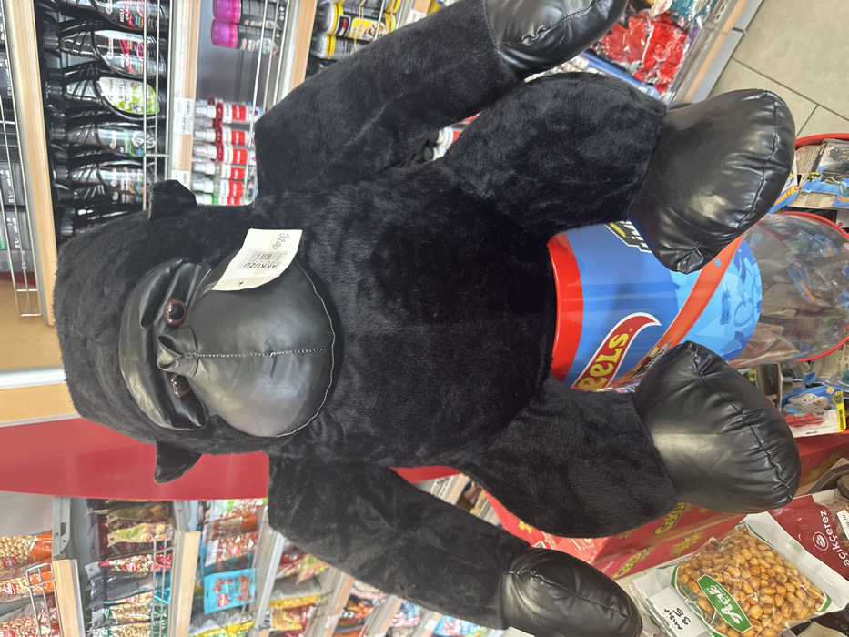 A large, black plush gorilla toy is prominently displayed within a retail store in Torbalı, Turkey. The gorilla, covered in black faux fur with black leathery material on its face, ears, hands, and feet, sits upright. It rests upon a cylindrical, transparent display bin featuring a red top rim and "Hot Wheels" branding, with small items visible inside. A white retail tag, displaying "AKKUZU" and "32.00", is attached to the gorilla's chest. The background features metal shelving units stocked with merchandise. To the left, numerous packaged food products, including bags of nuts (one partially visible with Turkish text 'Mısır 35'), line the shelves. To the right, shelves hold various bottles and aerosol spray cans. The scene is brightly lit, indicative of an indoor commercial environment. No individuals are visible.