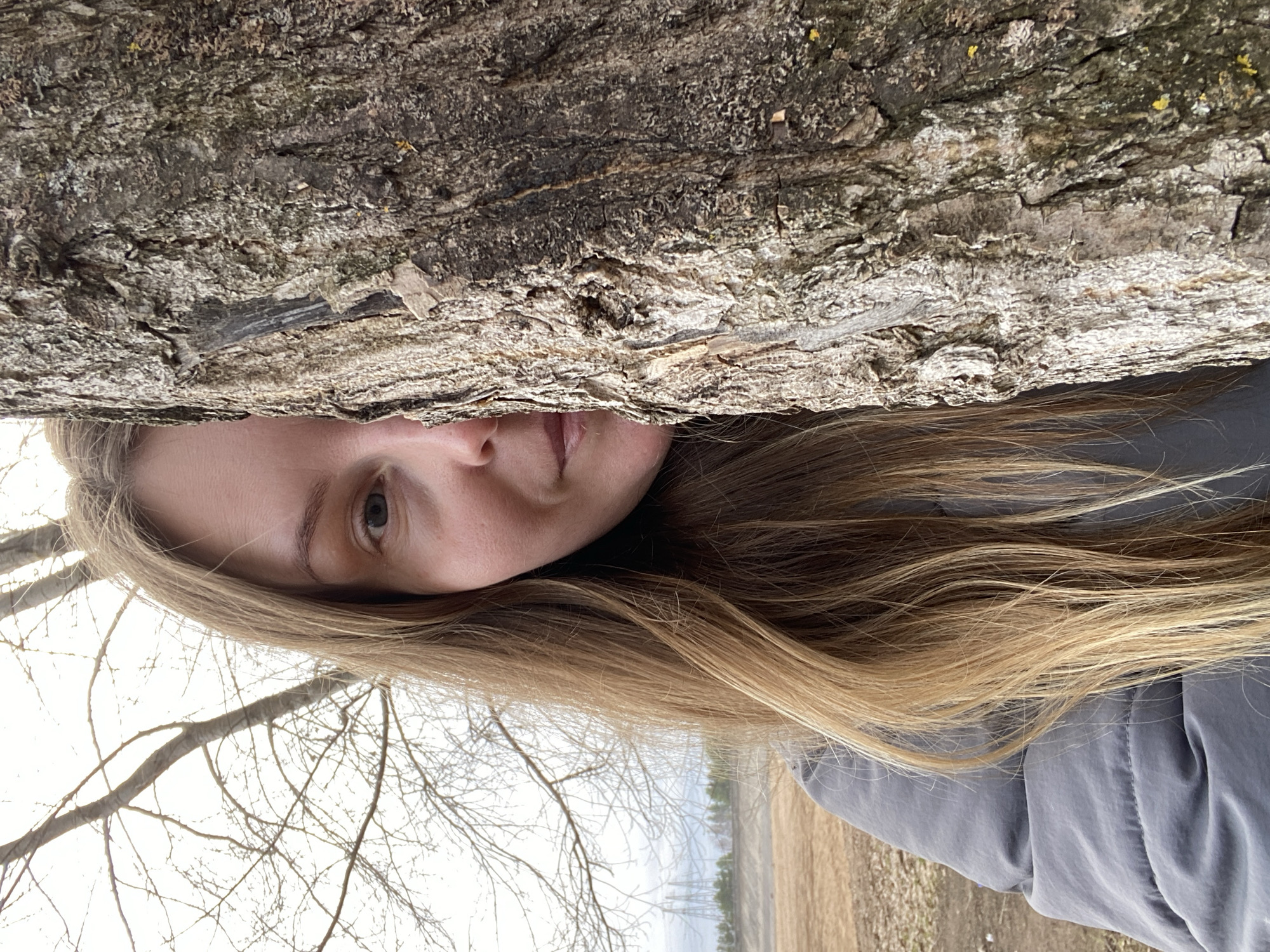 A woman with blonde hair is positioned beneath a tree trunk with rough bark. Her face is partially visible; she appears to be outdoors, likely in an area with trees, branches, and a field in the background. The location cannot be confirmed to be Noviy, Russia based solely on the provided image.