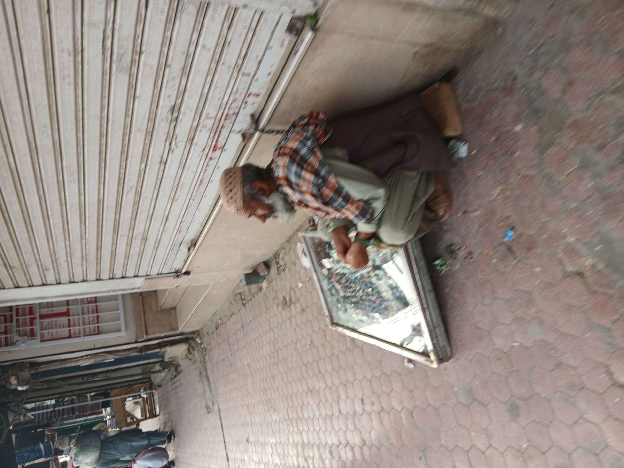 An elderly man with a white beard and a patterned hat is sitting on the ground next to a glass display case filled with various small items. He is wearing a plaid shirt and light green pants. The man is located on a brick-paved sidewalk in front of a closed storefront with a corrugated metal door. The scene is in Quetta City Tehsil, Pakistan.