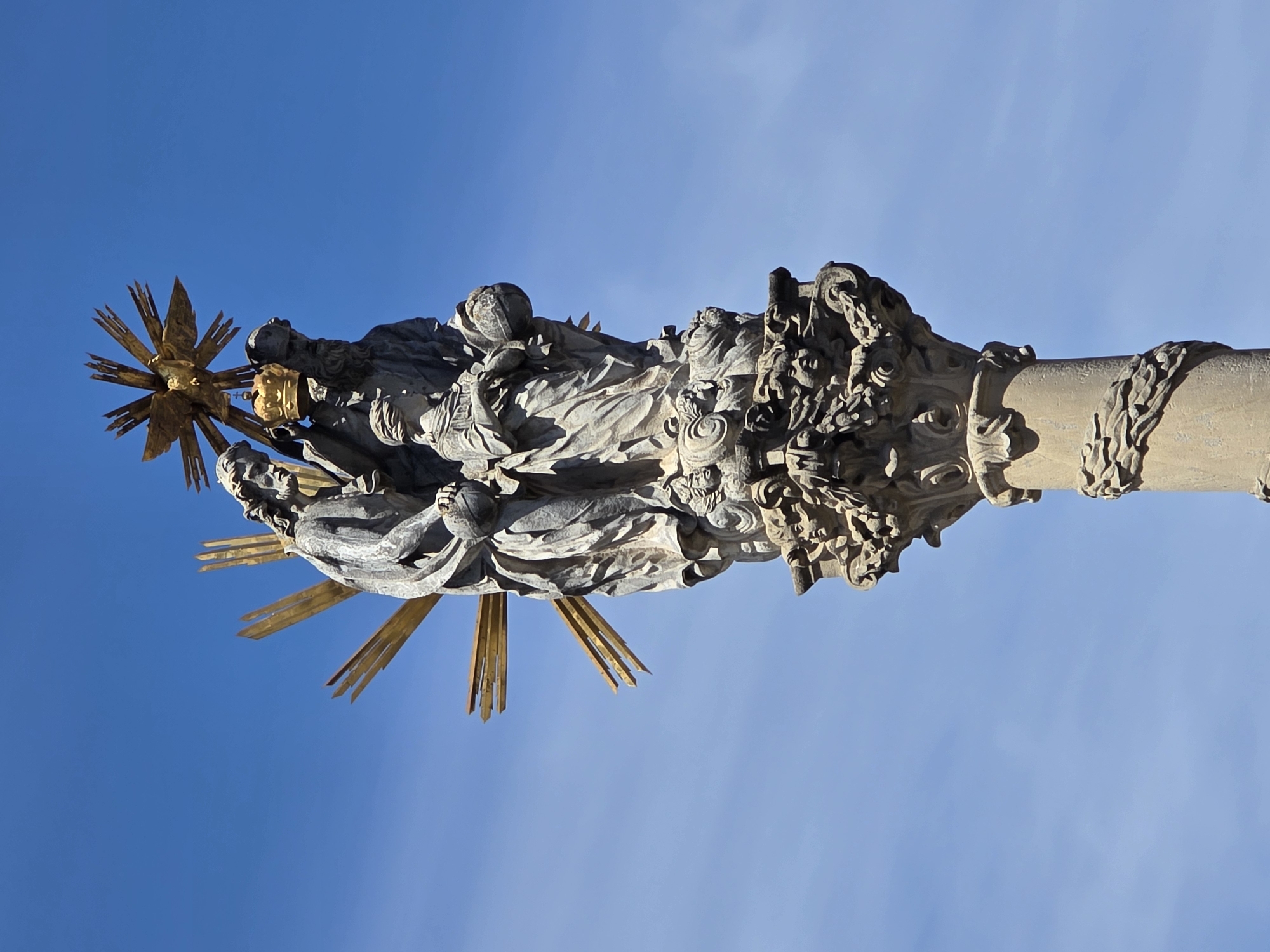 The image shows the Holy Trinity Column in the city of Trnava, Slovakia. It is a baroque plague column topped with a sculpture of the Holy Trinity, with God the Father, Jesus Christ, and the Holy Spirit represented as a dove. The column is made of stone and is adorned with intricate carvings, including cherubs and floral motifs. In the original image, the sky is blue and clear, indicating daytime. The column is viewed from below, emphasizing its height and grandeur. The sculpture is detailed, with each figure rendered in classical style. The Holy Spirit is depicted as a golden dove radiating light, surrounded by golden rays. Jesus is shown with a beard and wearing a crown, holding a globe. God the Father is also bearded and is depicted as an older man. The overall impression is one of solemnity and religious devotion.