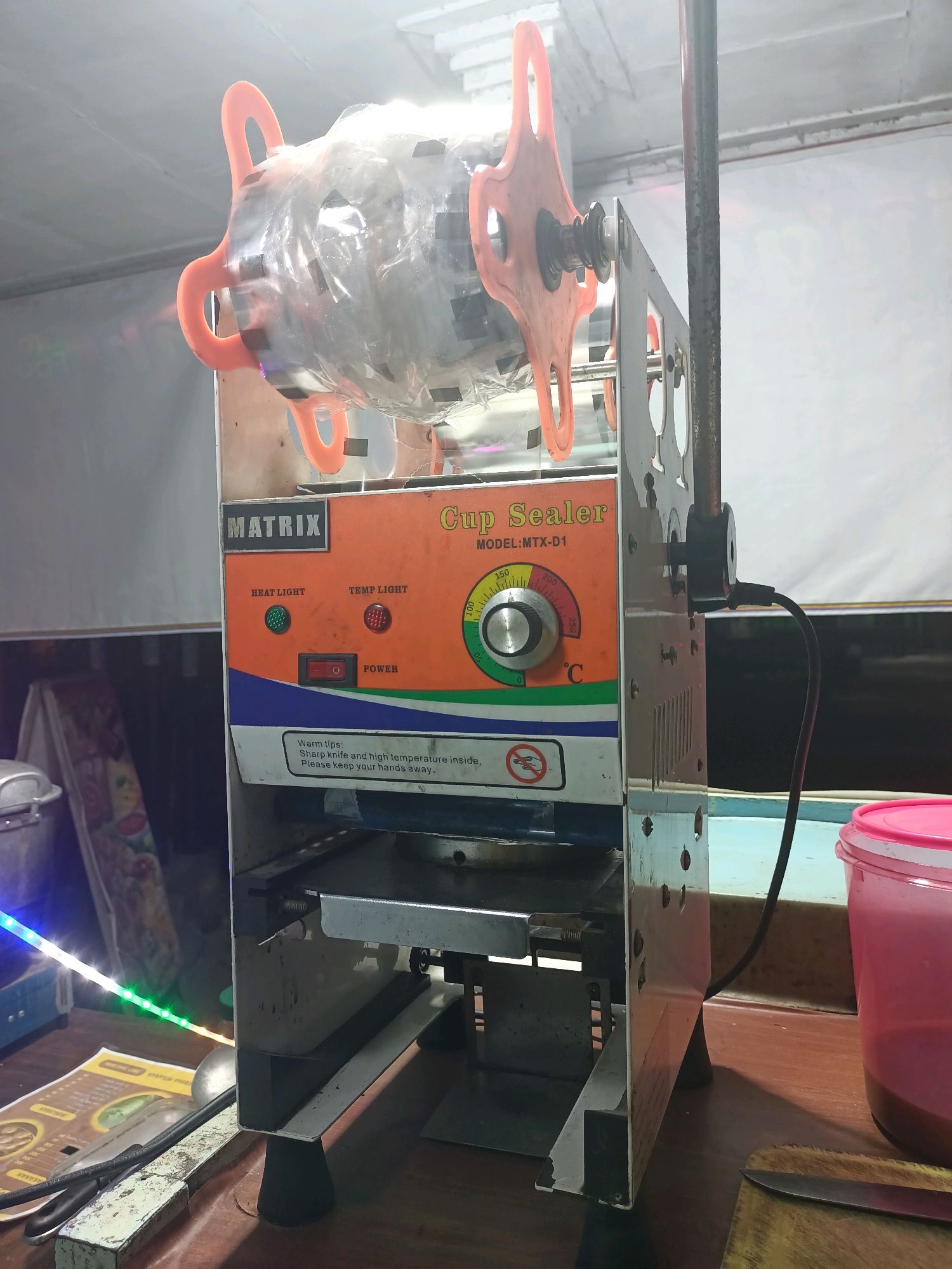 The image shows a cup sealing machine in what appears to be a small food stall or shop in Badas, Indonesia. The machine is orange and silver, with a roll of plastic film at the top. There are various controls and indicators on the front of the machine, including a temperature dial, power switch, and indicator lights. A warning message about sharp knives and high temperatures is also visible. In the background, there are shelves with some items and colorful LED strip lights. On the table in front of the machine, there are some papers, a ladle, a knife, and a pink plastic container. The overall scene suggests a functional, busy workspace.