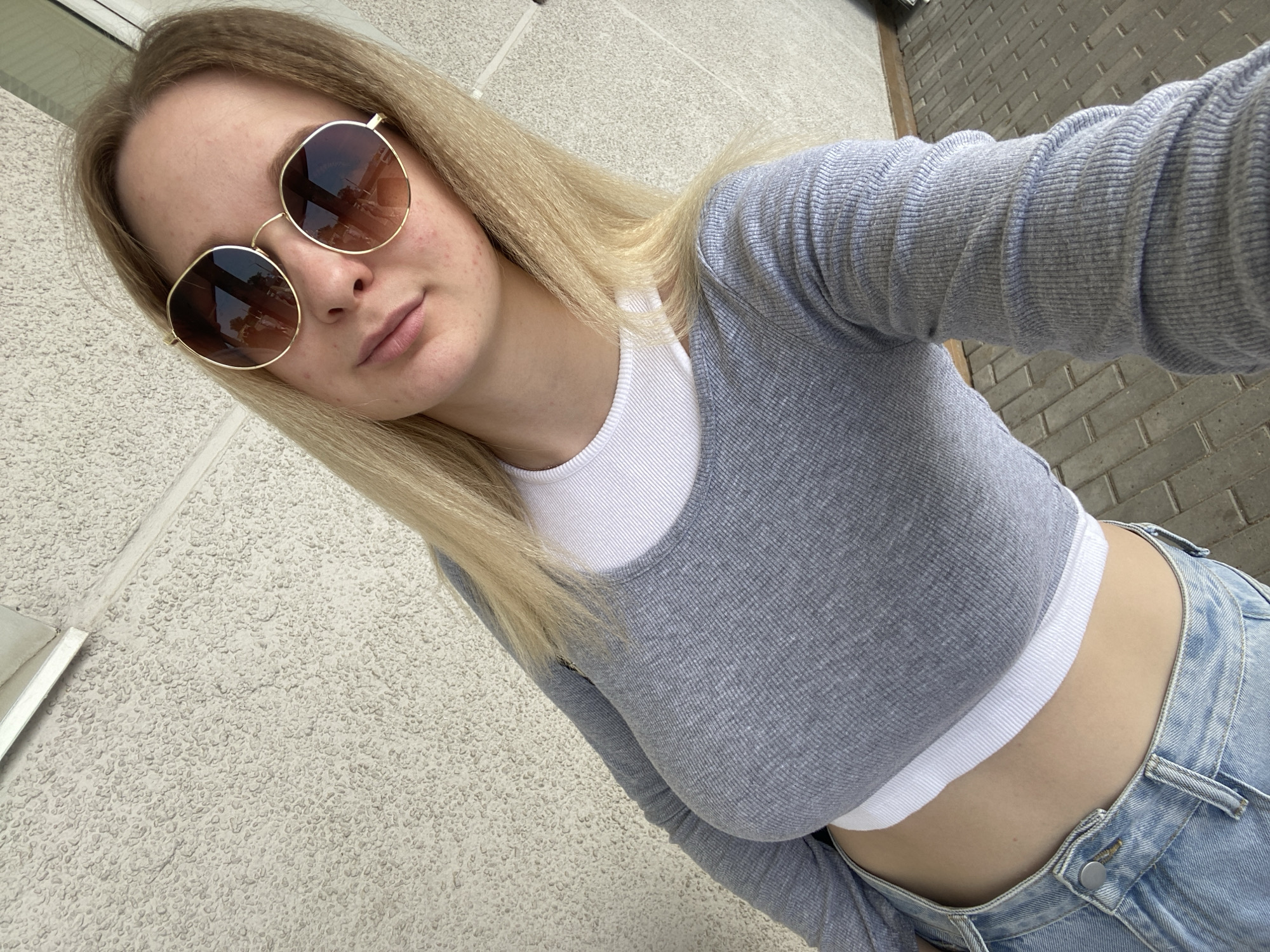 In a medium outdoor shot, a young Caucasian woman stands facing the camera. She has long blonde hair, and is wearing large, round, gold-rimmed sunglasses with brown lenses. She is also wearing a grey, long-sleeved crop top layered over a white tank top, and blue denim jeans. The background is a textured, light-colored wall. The ground appears to be a paved surface with rectangular stones. The lighting suggests it is daytime. The image is taken from a slightly elevated angle.