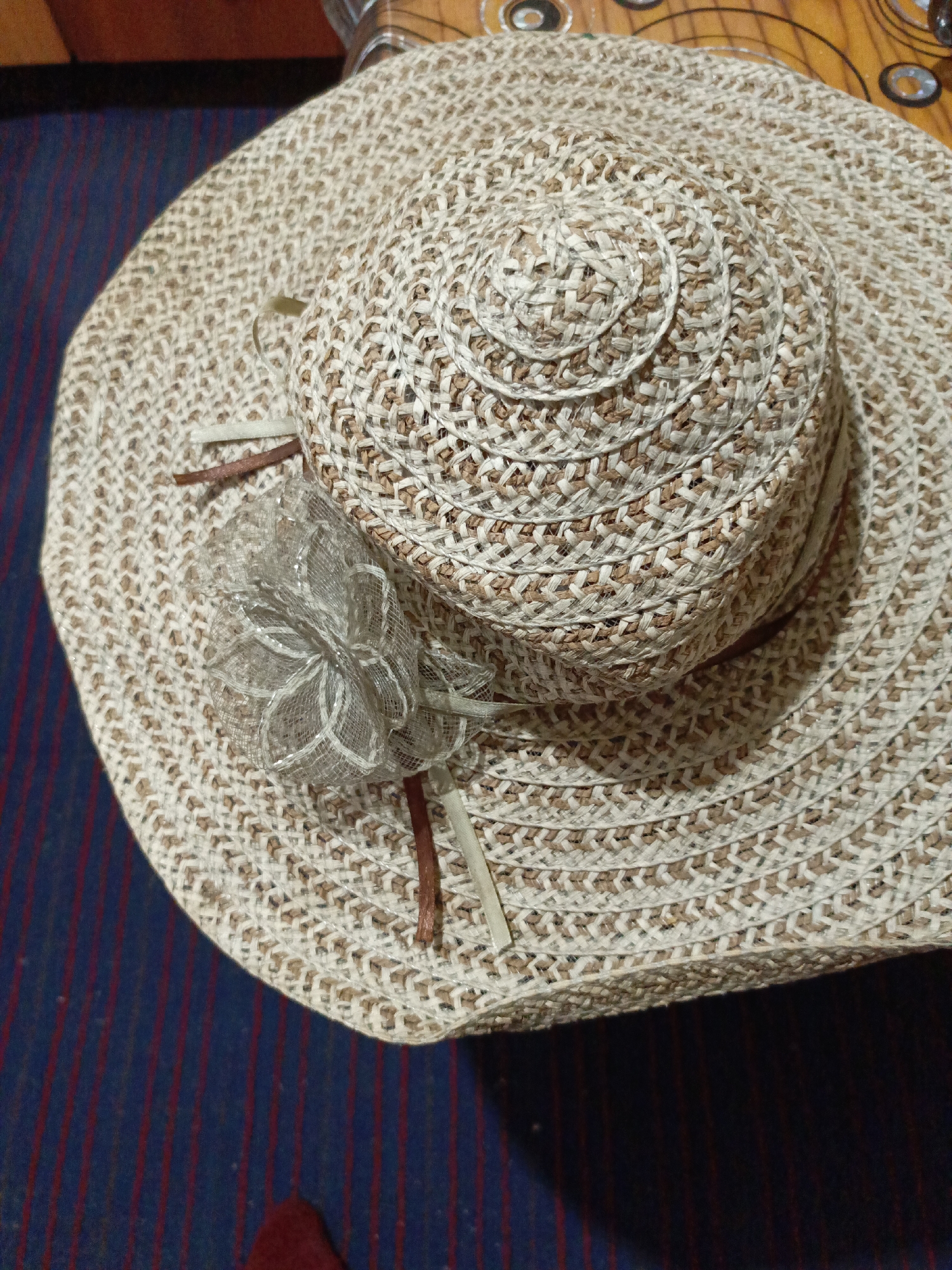 A woven sun hat is displayed indoors. The hat has a light tan and brown color scheme with a decorative netting flower and ribbon. It sits on a surface with blue and red stripes. The setting appears to be in a home. There is no information to confirm that the image was taken in Zalesovo, Russia.