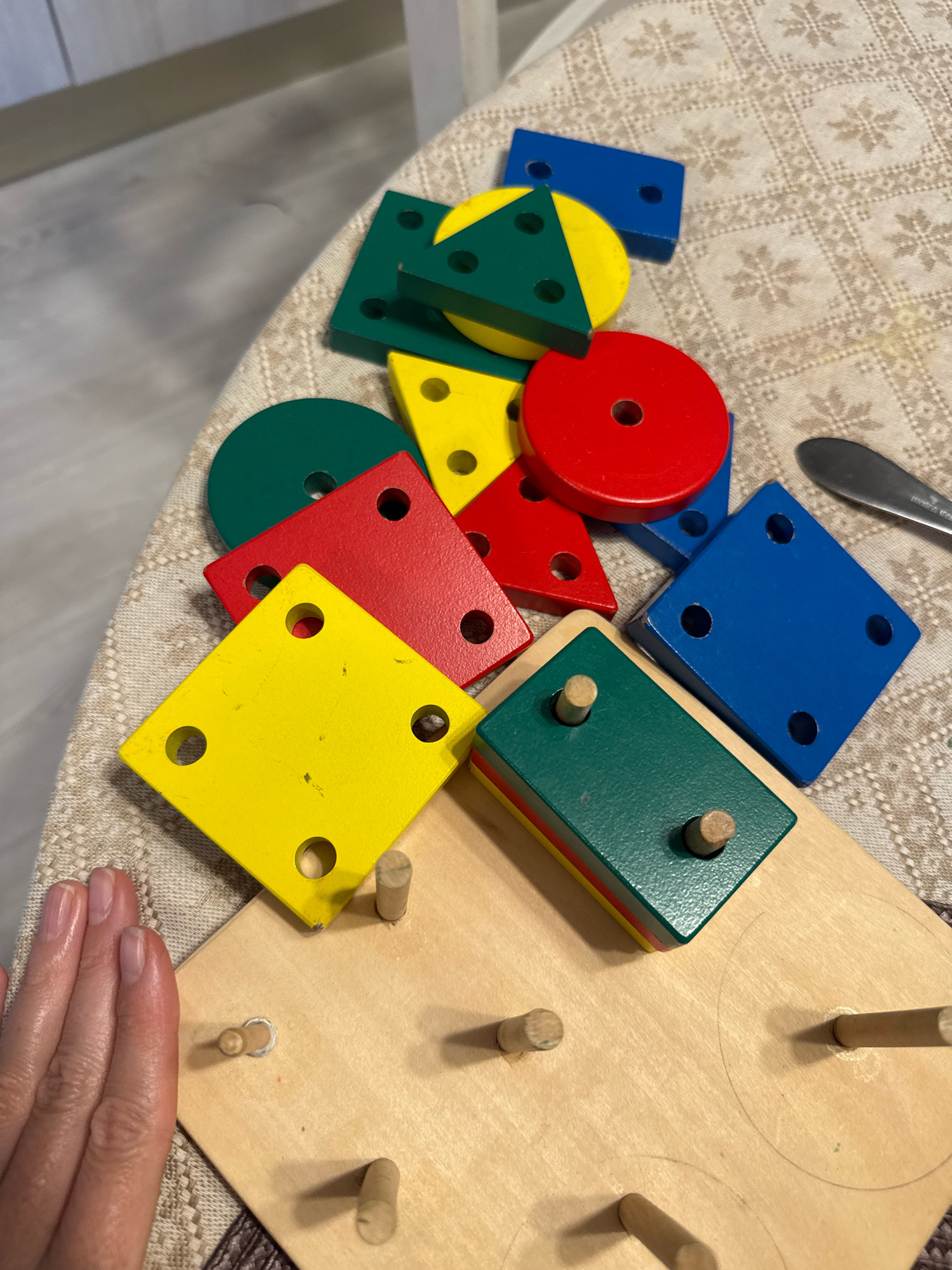 A top-down, medium shot captures a collection of colorful wooden geometric shape toys scattered across a patterned tablecloth. The toys include red, yellow, green, and blue squares, a red circle, a yellow triangle with green and blue segments, and a green rectangular block with red and yellow stripes. Several of these shapes have holes for pegs. The toys are arranged in a somewhat haphazard pile. In the foreground, a portion of a wooden sorting board with several pegs is visible. A person's left hand, with fingernails visible, is placed on the left side of the sorting board. To the right of the scattered shapes, a dark utensil, possibly a knife, rests on the tablecloth. The background is blurred, showing a light-colored wall and possibly a cabinet. The image does not provide any information about the location in Saint Petersburg, Russia.
