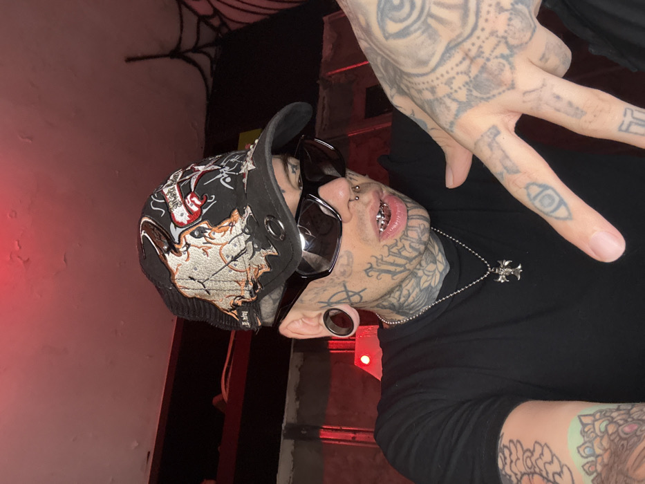 An individual, likely an adult male, is depicted in a medium-close-up, eye-level shot, looking directly at the camera. He wears a distressed black mesh-back baseball cap featuring an embroidered skull-like design and text. Dark, rectangular sunglasses obscure his eyes. His face, neck, and visible skin are extensively covered in tattoos, including an anarchy symbol on the left cheek, stylized lettering, and a lotus flower design on the neck. He presents large ear gauges in both earlobes, multiple nose piercings (two studs and a septum ring), and silver-colored grillz covering his upper teeth. He is dressed in a plain black short-sleeved t-shirt and wears a silver-colored ball-chain necklace with a detailed cross pendant. His arms and hands are also heavily tattooed, with the right hand raised and fingers slightly splayed, revealing an eye symbol on the index finger. The environment is dimly lit by a prominent red light source from above, illuminating a peeling white ceiling and casting a red hue on the scene. The background is dark and indistinct, featuring unidentifiable dark structural elements to the left and a white spiderweb decoration on a dark surface to the right. The specific location within the city of Buenos Aires, Argentina, cannot be determined from the visual information provided.