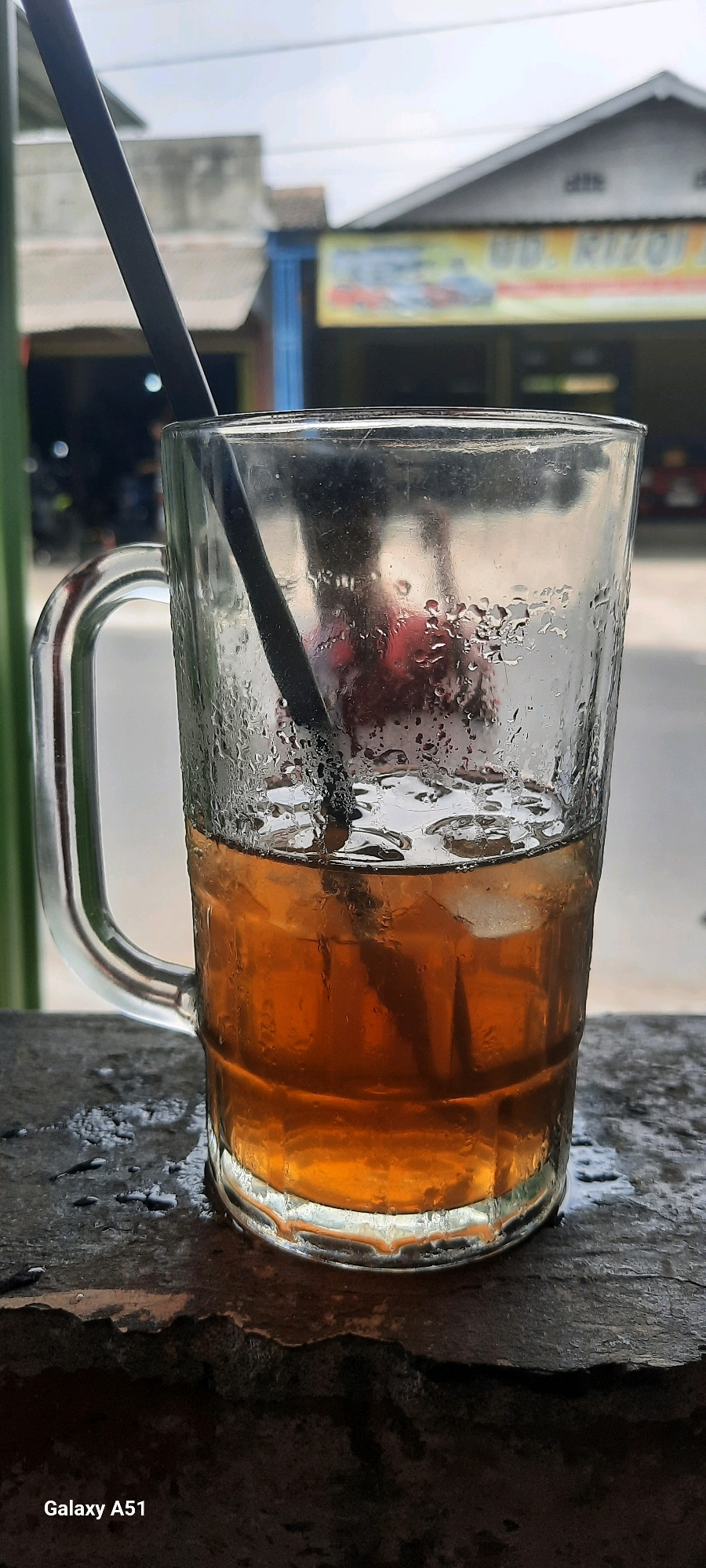 A clear glass mug filled with iced tea and ice cubes sits on a dark, textured surface. A black straw is submerged in the beverage. The mug's exterior is covered in condensation, creating water droplets. In the background, a blurred scene shows a building with a sign that reads "UD. RIZQI A." and what appears to be a street with some vehicles. The sky above is overcast. The location is identified as Selopuro, Indonesia, indicated by the text "Galaxy A51" in the bottom left corner, a common watermark for Samsung phones, which are widely used in Indonesia.
