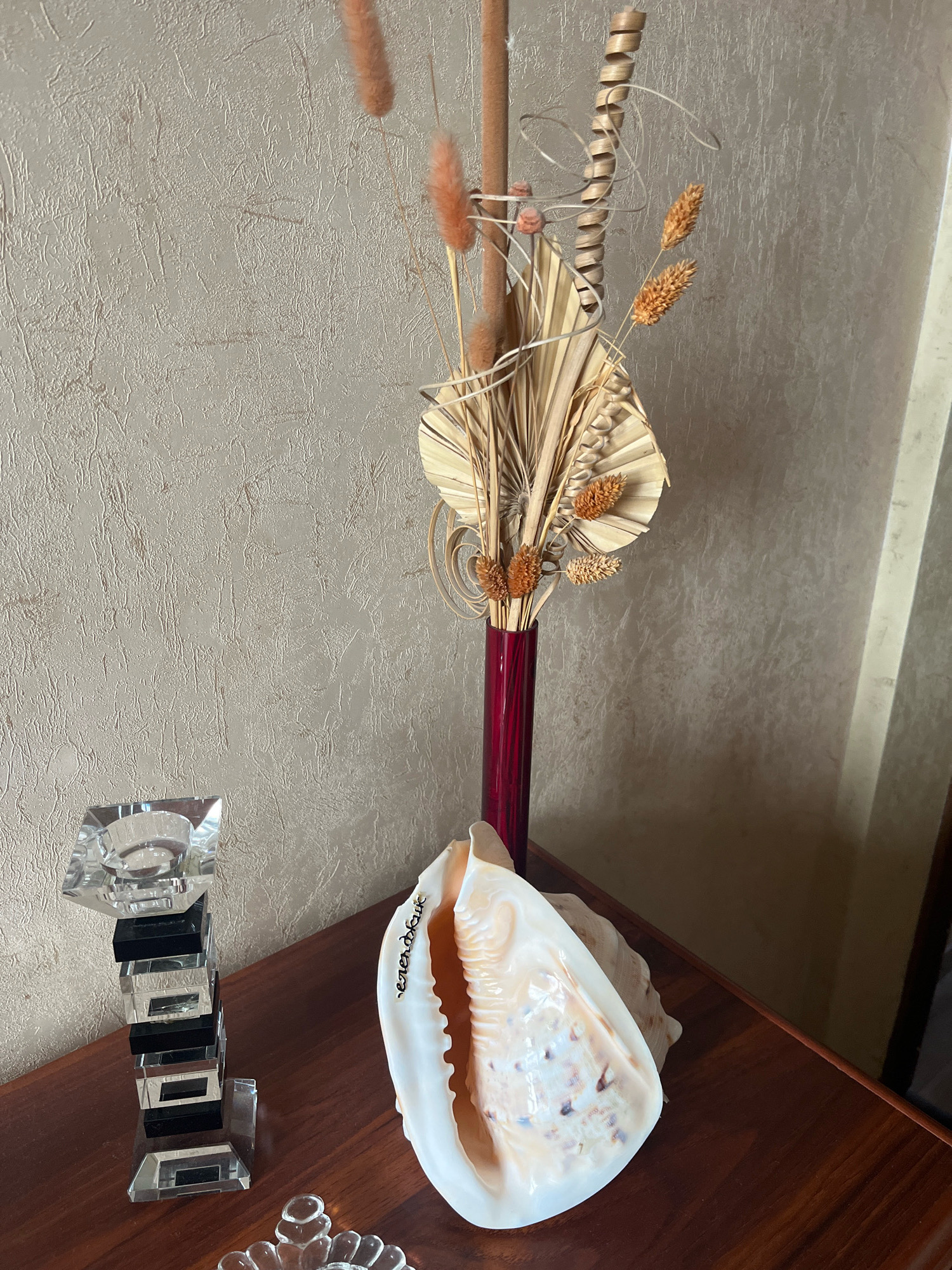 The image depicts a still life arrangement on a dark wooden surface, likely a table or shelf, against a textured beige wall. The primary objects are a large, decorative seashell positioned prominently in the foreground, a tall, slender red vase containing dried decorative flora, and a crystal and black rectangular candlestick to the left. The seashell is open, revealing its intricate inner texture and a pearlescent sheen, with some dark speckles. On the interior surface of the shell, there appears to be faint, handwritten text in a light color. The dried flora in the vase includes palm fronds, feathery stalks in shades of brown and tan, and a spiral decorative element. The candlestick is made of clear, faceted crystal and black rectangular inserts, stacked vertically to create a stepped effect. A small, clear decorative object with a floral design is partially visible at the bottom center of the frame. The lighting suggests an indoor setting with ambient light casting shadows. The image does not contain any individuals or indicate any activity. There is no information within the image to suggest a location in Vladimirovka, Russia.