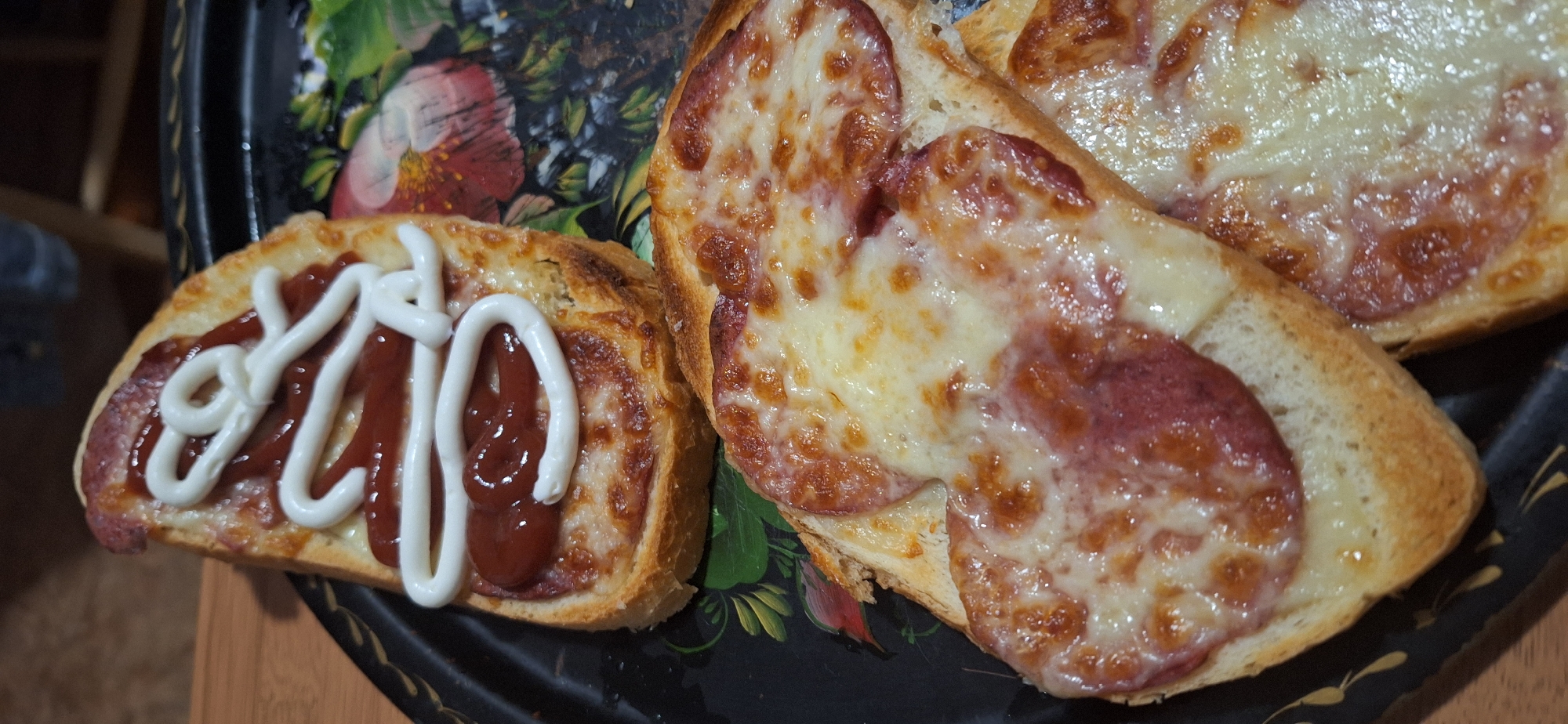The image depicts three pieces of toasted bread, two topped with melted cheese and sliced sausage, and one with sliced sausage covered in ketchup and mayonnaise. They are arranged on a decorative serving tray with floral patterns. The background is blurred, but the tray rests on a wooden surface. There is no indication of a specific location in Blagoevgrad, Bulgaria, as the image does not contain any landmarks or identifiable features related to that city. The scene is focused on the prepared food items.