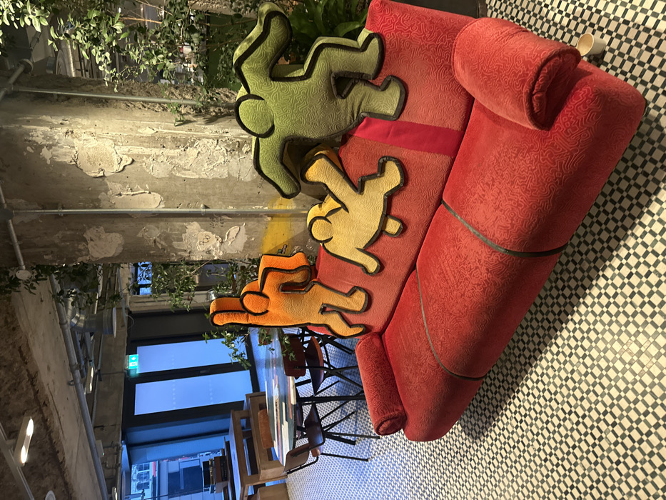 A red patterned sofa sits on a black-and-white tiled floor. The sofa has three human-shaped cushions in descending order from top to bottom: green, yellow, and orange. The cushions are stylized with a black outline. The setting appears to be a cafe or lounge, with tables and chairs visible in the background through a window. Exposed pipes and foliage decorate the space, providing an industrial yet organic feel. The image's orientation is rotated clockwise.
