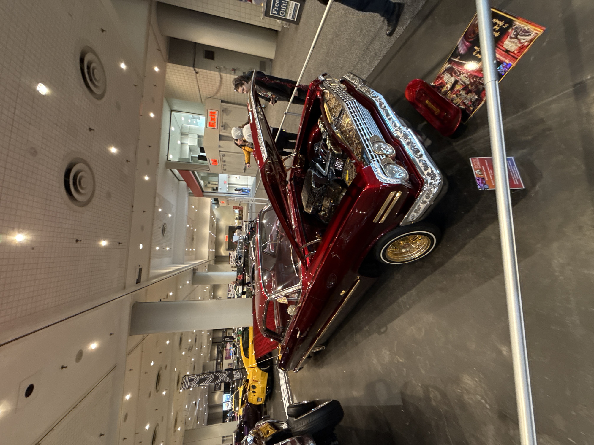 The image shows a red classic car on display with its hood open, featuring custom chrome detailing and gold rims. The car is likely part of a car show or exhibition. Several other vehicles, including a bright yellow car, can be seen in the background, suggesting a display area with multiple cars.
People can be seen near the car, some of whom appear to be admiring or observing the vehicles on display.
The location appears to be a large indoor event space. The presence of multiple cars indicates a professional event setup. The environment is well-lit, with a focus on highlighting the exhibited cars.
The image was likely taken in the city of New York, United States of America. The distinctive style and type of cars are associated with car customization scenes often found in urban areas. The scale of the event suggests a major city venue.