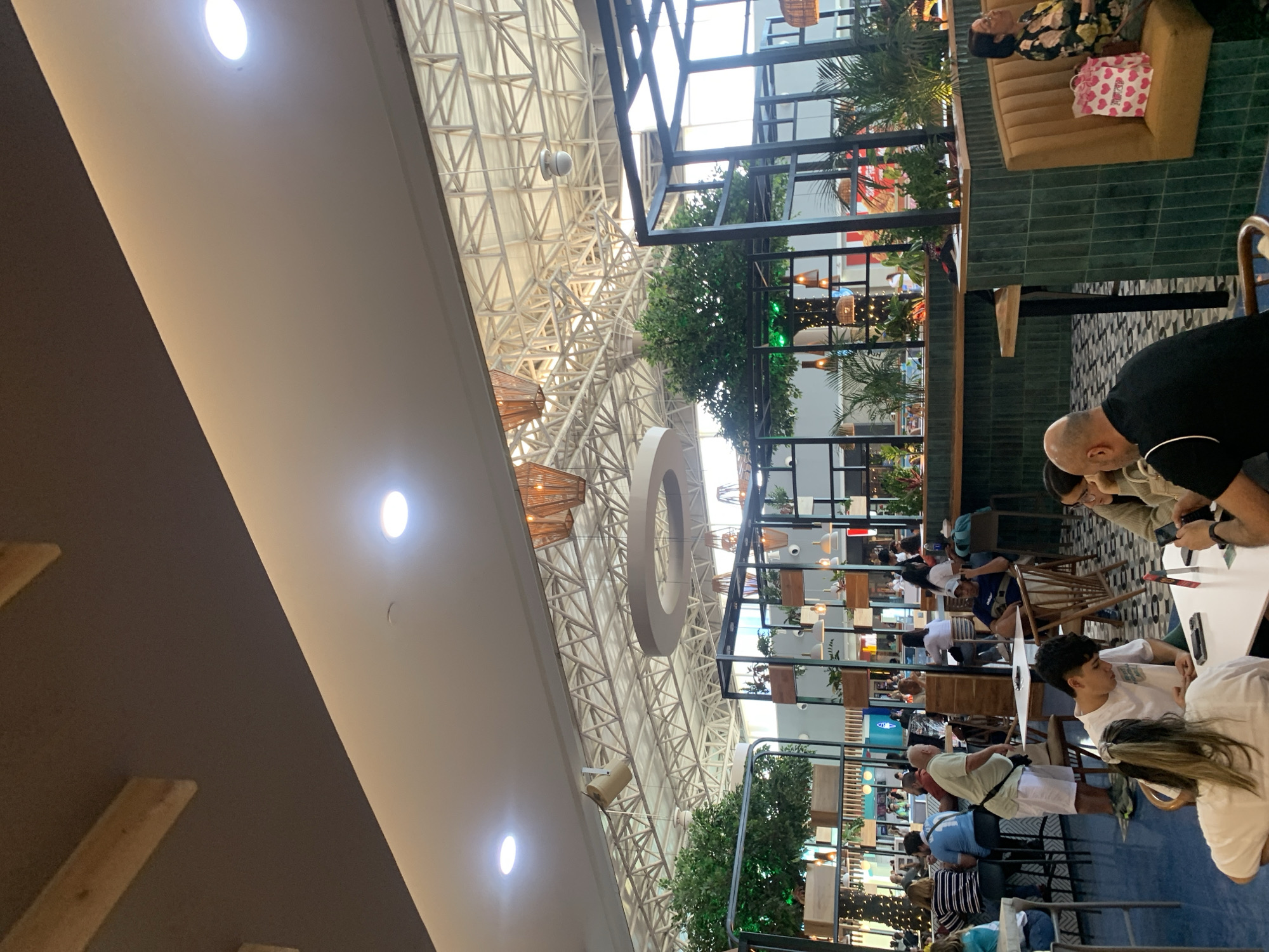 This image depicts the interior of a commercial space, likely a restaurant or mall food court in Porlamar, Venezuela. The focus is on the ceiling, featuring a complex network of white structural beams and lighting fixtures, including circular drop pendants. Below, there are tables with people seated, some appearing to be engaged in dining and conversation. The foreground shows individuals seated at tables, some interacting with electronic devices. In the background, there's a multi-level structure with plants and possible retail or food service vendors. The scene overall suggests a modern indoor public space.