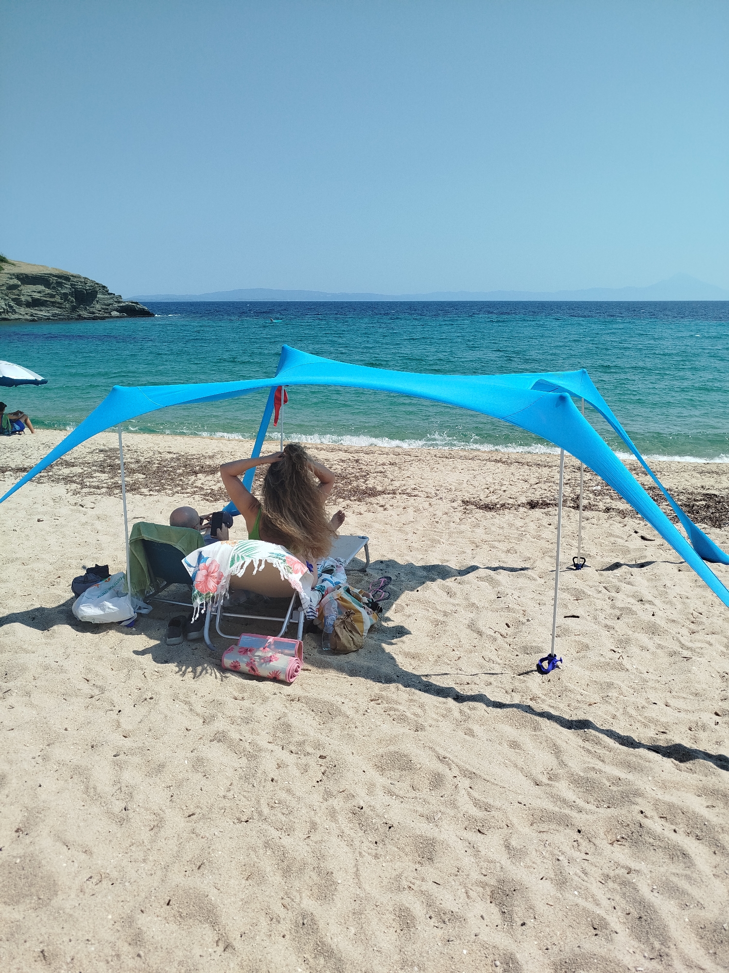 On a sandy beach in Sithonia Municipal Unit, Greece, two individuals are seeking shade under a blue beach tent. The tent is supported by poles anchored to the sand. The sea, a vibrant turquoise, is visible behind them, with a gentle wave line breaking on the shore. A rocky outcrop juts into the water on the left side of the frame. Scattered on the sand around the tent are various beach items, including chairs, bags, and towels. One person, with long, light brown hair, is seated, facing away from the camera, and appears to be adjusting their hair. The other person is seated in a chair to the left, also with their back to the camera, possibly looking at their phone. The sky is a clear, bright blue.