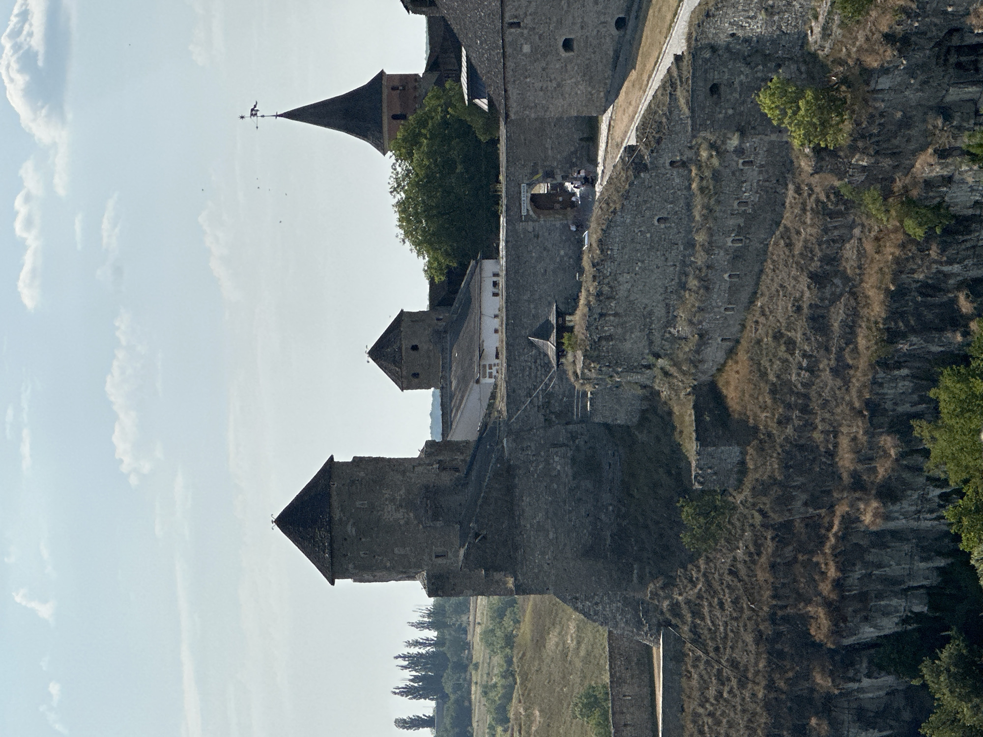The image shows a scenic view of the Kamianets-Podilskyi Castle in Ukraine. The ancient fortress, constructed from grey stone, stands prominently on a rocky cliff overlooking a landscape of dry grass and scattered trees. The castle complex features several towers, including a tall, cylindrical tower with a dark, conical roof on the left, and a taller, narrower tower with a pointed roof and a weather vane on the right, which appears to be part of a church. In the center, a lower, square building with a dark roof connects to other fortified walls. An arched gateway, likely an entrance to the castle grounds, is visible in the middle of the image, with several figures walking through it. The sky above is a light blue with scattered white clouds, and distant trees form a line on the horizon. The overall impression is one of historical grandeur and resilience.