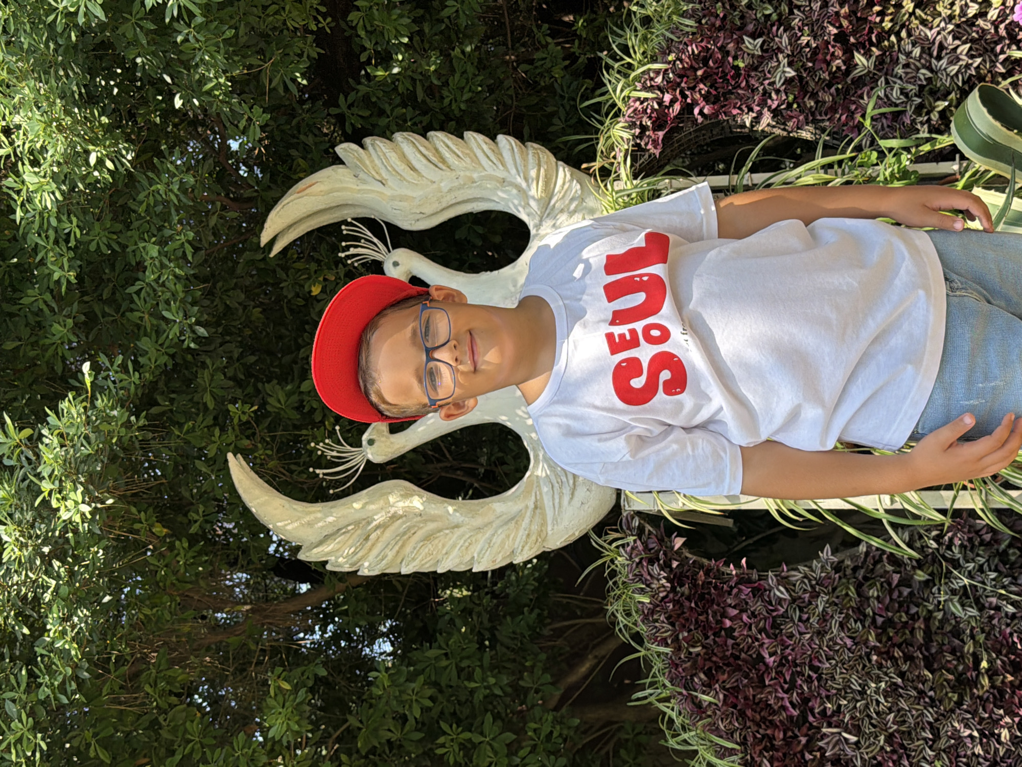 A young boy wearing a white t-shirt with red lettering, blue-rimmed glasses, and a red baseball cap stands in front of a white sculpture of two birds with wings spread. He is in front of a lush green tree canopy and a wall of purple and green foliage. The boy is smiling. The image appears to have been taken outdoors during the daytime. There are no discernible landmarks to suggest the location is in Sochi, Russia.