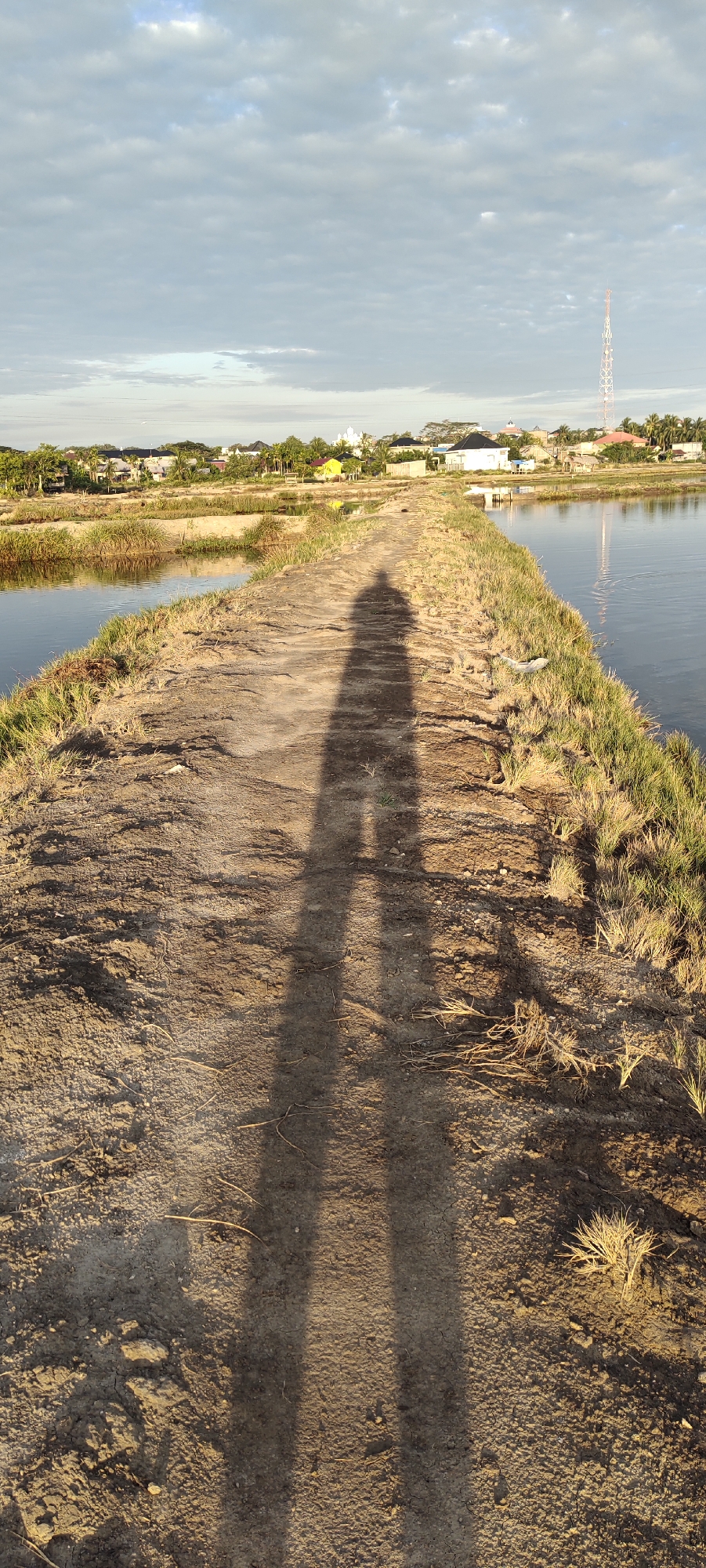 The image captures a dirt path flanked by bodies of water, likely aquaculture ponds, in Lhokseumawe, Indonesia. The path is covered in dry soil and scattered dry grass. The shadow of two individuals, presumably standing together, stretches prominently across the path, indicating the sun is low in the sky, likely during sunrise or sunset. In the background, a residential area with houses and buildings is visible, along with a tall communication tower. The sky is partly cloudy, with the sun casting a warm, golden light. The overall scene depicts a quiet, rural or semi-rural setting.