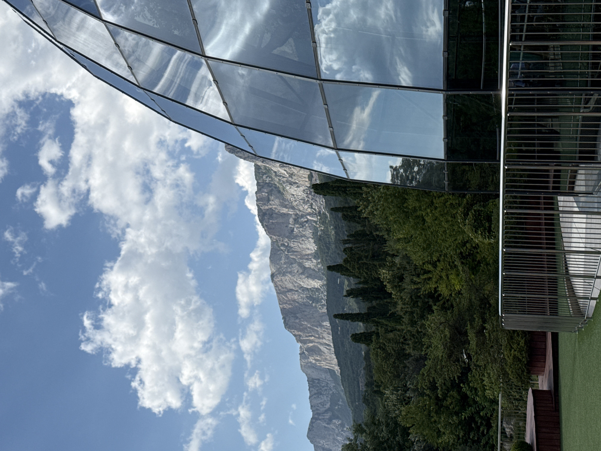 The image captures a scenic view from the city of Simeiz, Ukraine, featuring a striking modern glass-domed structure in the foreground. The structure's multifaceted glass panels reflect the bright blue sky dotted with fluffy white clouds, as well as the lush green trees and the imposing, rocky mountain range that forms the backdrop. In the middle ground, a dense forest of various trees, including tall, dark cypresses, transitions into the lower slopes of the mountains. A metal railing runs along the edge of a grassy area, suggesting a public or visitor space. The overall scene is one of harmonious coexistence between contemporary architecture and the natural beauty of the Crimean landscape.