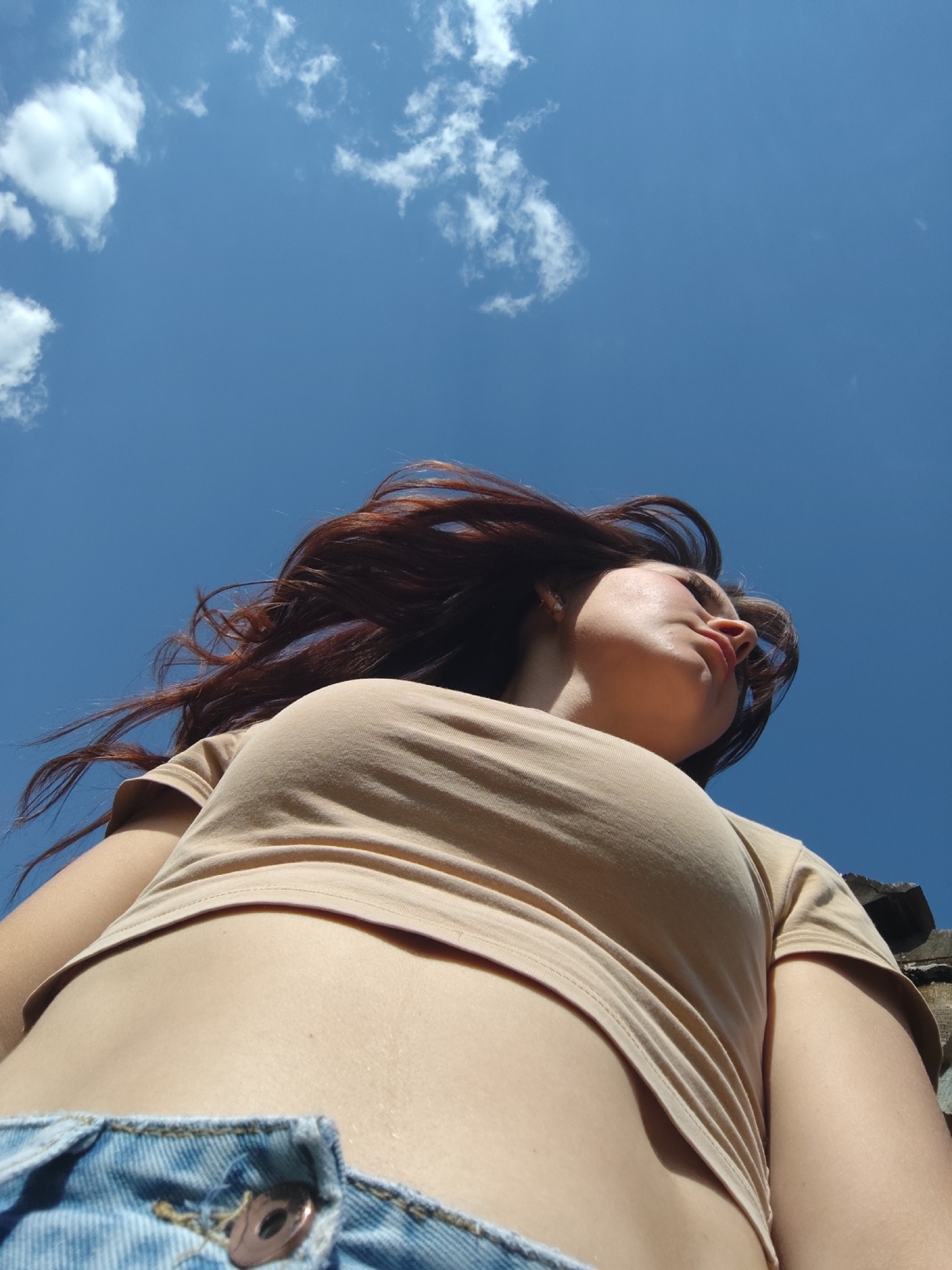 A low-angle, full outdoor shot features a woman with long, auburn hair lying on her back. She is wearing a beige, short-sleeved crop top and denim jeans, with the top button visible. Above her is a bright blue sky scattered with wispy white clouds. The focus is primarily on the woman's torso and the sky. There are a few blurry shapes on the lower right corner. The location in the city of Фоки, Russia cannot be confirmed, as the image does not contain any information about the location.