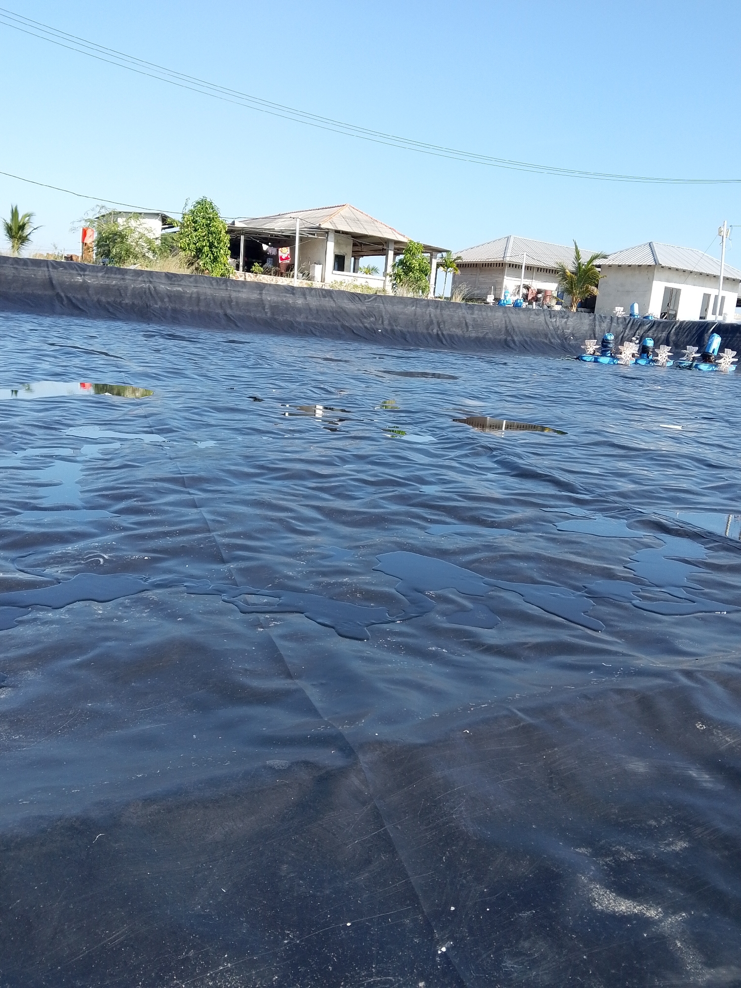 The photograph depicts a black, seemingly artificial pond or water containment area in Bukit Ketok, Indonesia. The body of water is lined with a black material, possibly a tarp or liner, which is slightly wrinkled and reflects the clear blue sky overhead. There are small pools of standing water on the surface of the liner. Along the edge of the pond are several buildings, including small houses with varying roof styles and a possible open-air structure. There is some vegetation including trees and shrubs visible. In the distance, there are several aeration devices placed in a row along the back edge of the pond, used for water treatment or aquaculture purposes. An overhead power line is visible against the sky. The overall impression is of a rural or semi-rural scene with a man-made body of water for some industrial or agricultural purpose.