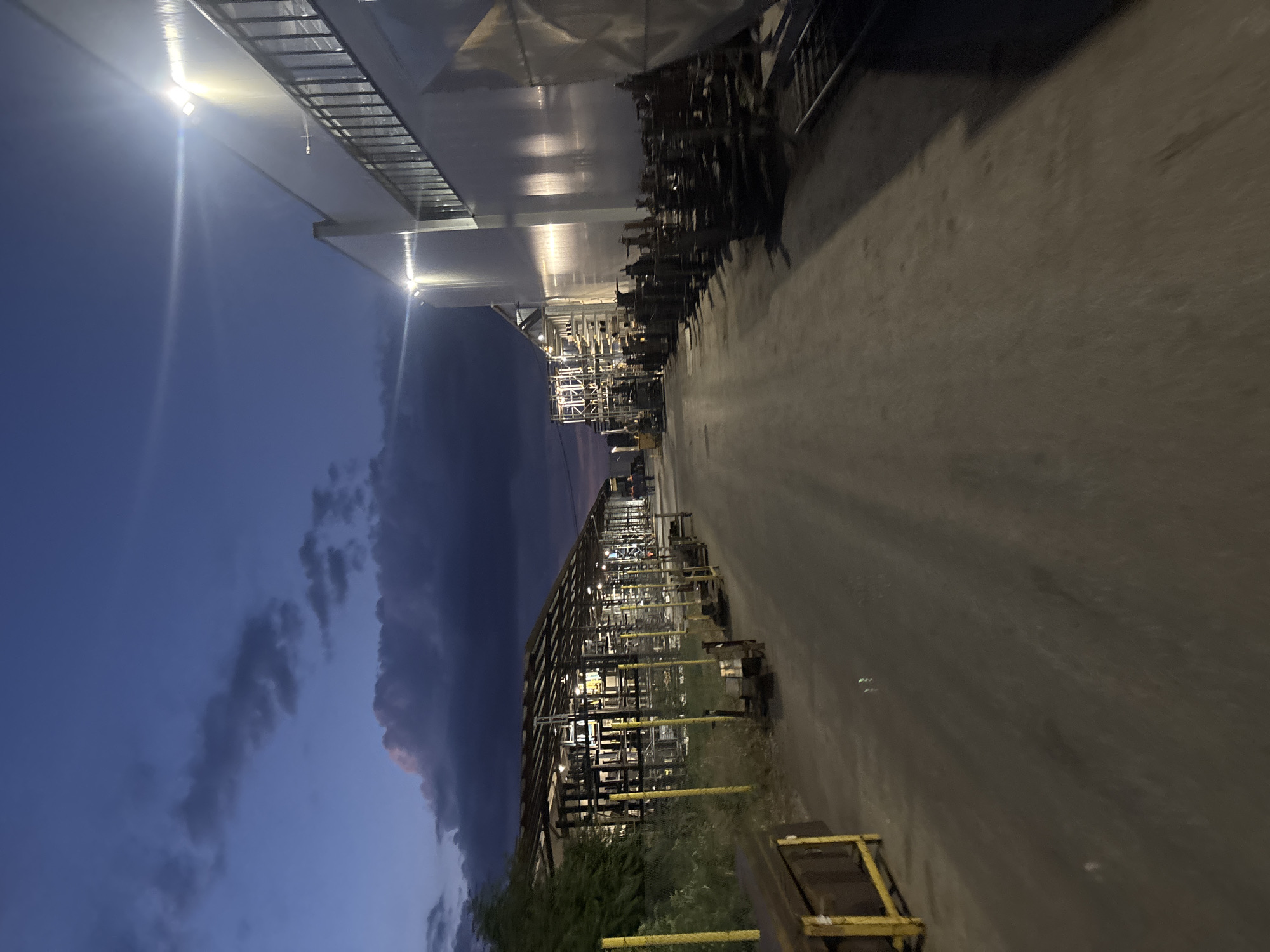 The image depicts an industrial facility at dusk. The sky is a deep blue with streaks of dark clouds. Two large, brightly lit buildings dominate the right side of the frame. Their facades are a pale, reflective material, and they are illuminated by powerful overhead lights that cast long shadows onto the paved ground. To the left, a covered open-air structure, likely for storage or manufacturing, is visible. It has a metal roof supported by a framework of vertical and horizontal beams. Rows of metal racks, possibly for materials, are lined up under this structure. In the foreground, a portion of a yellow metal container or storage unit is visible on the left. The ground is a dark, dusty asphalt or gravel surface. The scene appears to be devoid of significant human activity, with only a few indistinct figures visible in the distance on the left side of the frame near the covered structure. The overall impression is one of an operational industrial site during twilight hours. The location in Ruptsi, Bulgaria, is suggested by the context provided, though no specific geographical markers are present in the image itself.