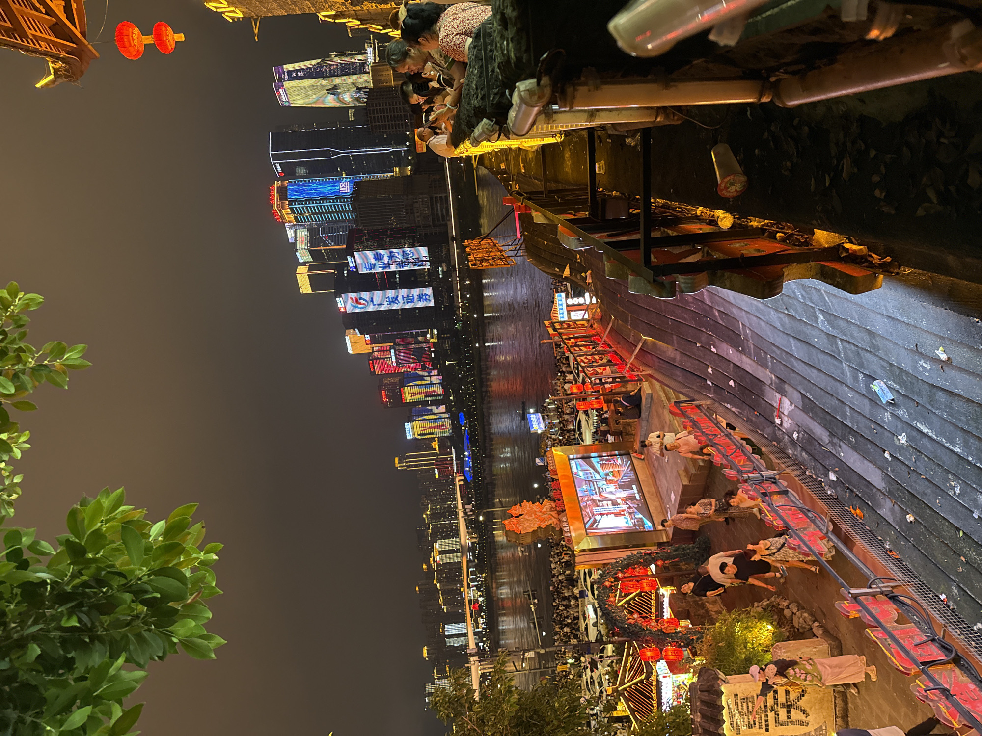 The image captures a vibrant night scene along the Yangtze River in Yuzhong District, Chongqing, China. In the foreground, a tiered walkway lined with red lanterns and ornate railings overlooks a bustling crowd gathered by the river. People are seen walking, standing, and interacting with each other. A large screen displays a video, drawing attention.
Across the river, the Chongqing skyline is illuminated by the colorful lights of numerous skyscrapers, creating a dazzling backdrop. The water reflects the city lights, adding to the dynamic ambiance. The overall atmosphere suggests a lively public space filled with both locals and tourists enjoying the evening. The scene is characterized by a mix of traditional Chinese architectural elements, such as the red lanterns and roof details, and modern urban development.