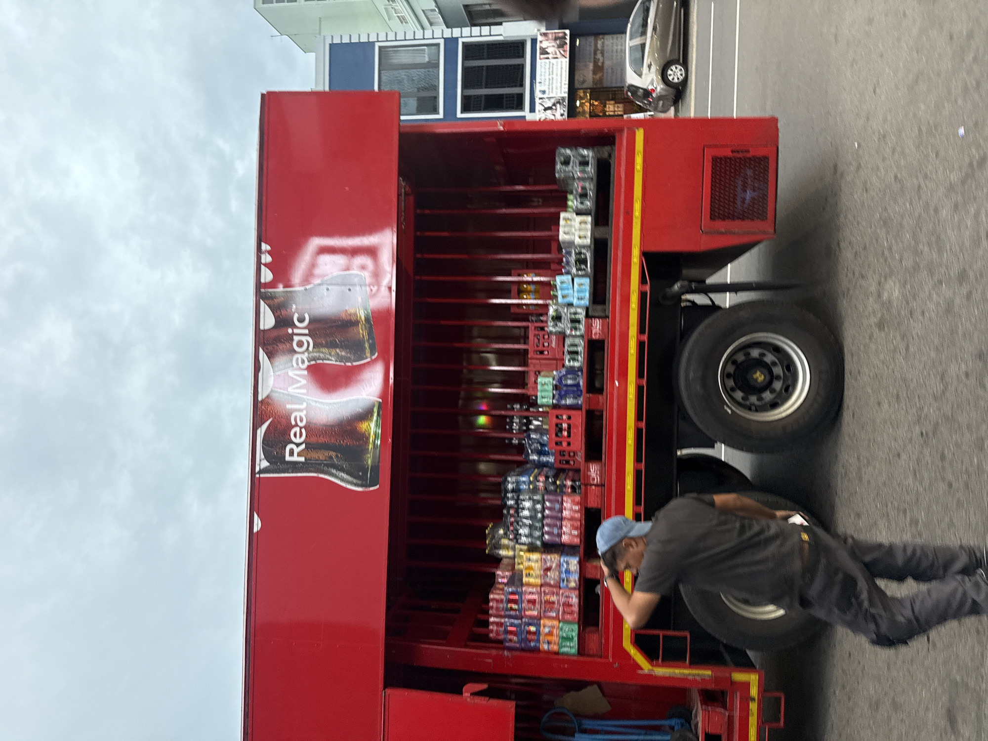 The image shows a red delivery truck parked on an asphalt road in Cape Town, South Africa. The side of the truck has "Real Magic" written on it. The truck's cargo area is open and filled with crates and packs of drinks. A man wearing a blue cap, gray shirt, and dark pants is standing near the rear of the truck. He is leaning towards the open cargo area, looking at a cell phone in his right hand. The background features buildings and a parked car. The sky is cloudy.