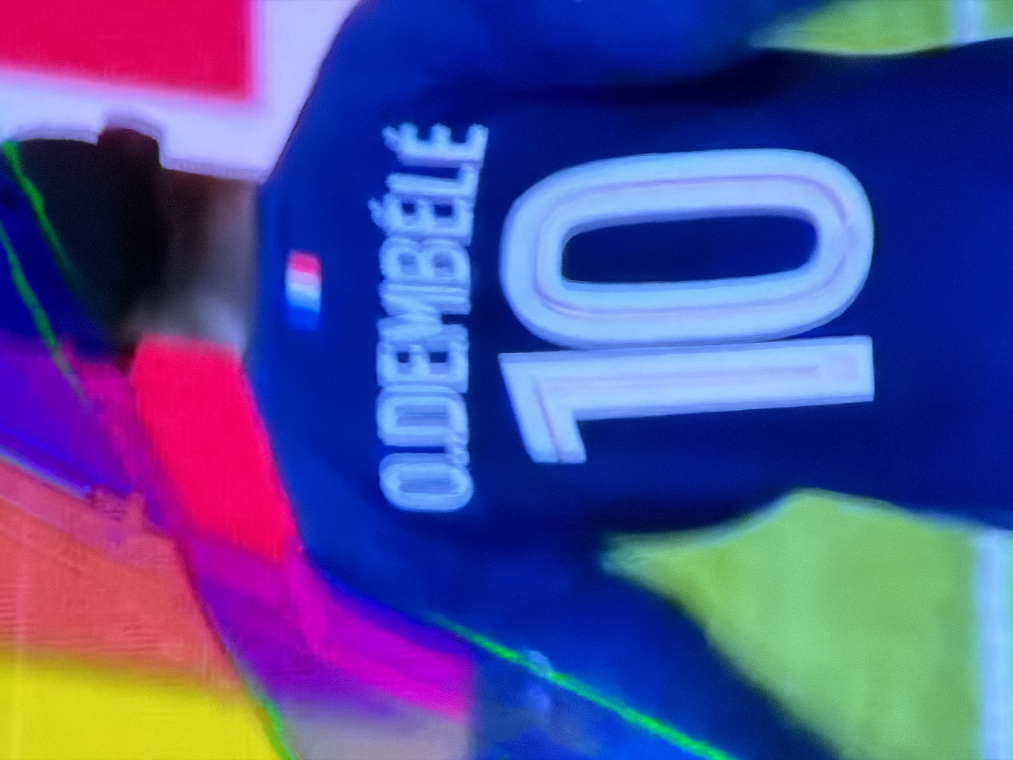 The image shows the back of a person wearing a blue jersey with the name "O. DIEMBELE" and the number "10" in white lettering. The French flag is visible on the jersey. The background appears to be a blurred mix of colors including red, pink, yellow, and green. The image is not in the city of Asenovgrad, Bulgaria.