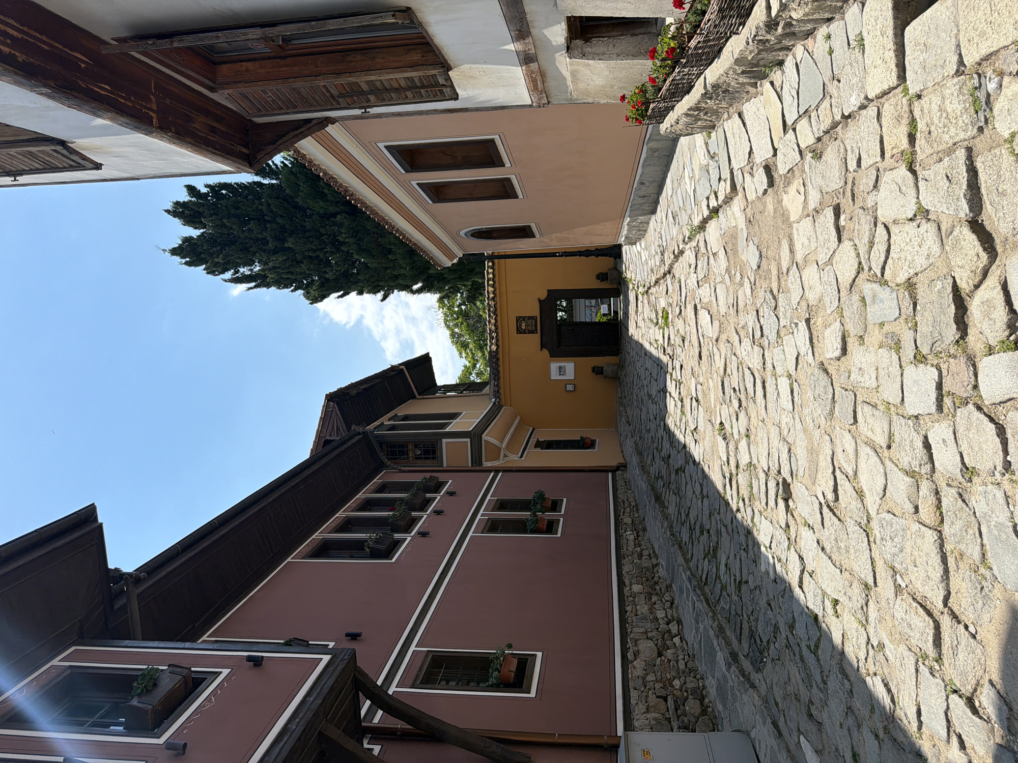 The image depicts a cobbled street within the Old Town of Plovdiv, Bulgaria, characterized by its distinctive National Revival period architecture. The perspective is longitudinal, looking down the street towards a further building and a tall tree under clear skies.
On the left side of the street, a two-story building with a deep reddish-brown (maroon) facade is prominent. Its exterior features white horizontal and vertical decorative bands. The upper floor exhibits a significant overhang, supported by dark wooden beams, with a dark brown roof and gutters visible at the top. Several windows, some with open wooden frames and others with dark wooden shutters, are visible. Green plants in planters are present beneath some of the windows. A light grey utility box is partially visible at the bottom left corner. Further along the street on the left, a section of another building, painted in a muted ochre or mustard yellow, is visible. This section includes a projecting bay window on the upper floor, also with dark wooden elements.
On the right side of the street, a two-story building with a pale peach or light beige facade is present. It also displays white decorative lines. Windows on this structure feature dark wooden frames and partially open wooden shutters. A decorative woven planter containing red flowers, likely geraniums, is situated below one of the lower-level windows. The base of this building is constructed from rough-hewn stone blocks. Further down the street on the right, another building with a similar pale facade features multiple windows, some arched.
The street itself is paved with irregularly shaped, grey and light brown cobblestones, interspersed with sparse green vegetation in the gaps. A distinct diagonal shadow line crosses the cobblestone surface, indicating direct sunlight from an off-center source.
At the visible end of the street, a bright yellow building with a dark, arched entrance and a mounted sign above is situated. Beyond this, a tall, dark green, coniferous tree, likely a cypress, stands prominently against the sky. A black lamppost is positioned near the yellow building.
The sky is bright blue with a few faint white cirrus clouds, particularly visible in the upper right quadrant. No individuals or vehicles are present in the scene.