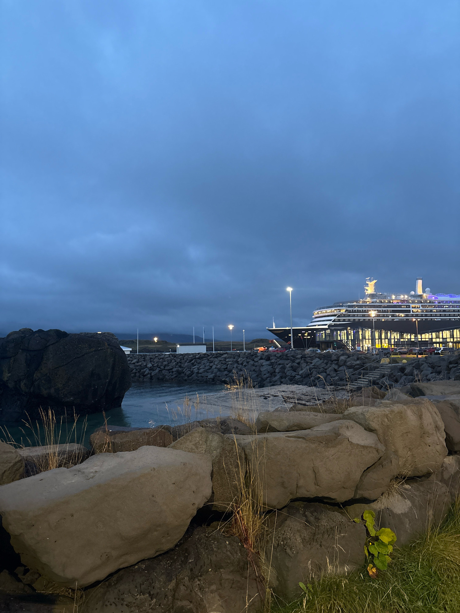 The image depicts a twilight scene at a port in Reykjavik, Iceland, with a large cruise ship docked at a terminal building. In the foreground, large grey rocks and sparse dry grass dominate the lower portion of the frame. To the left, a massive dark boulder sits partially submerged in the calm, dark blue water. In the mid-ground, a breakwater made of dark grey rocks separates the water from a paved area where several cars are parked. The cruise ship, illuminated by numerous lights, is positioned to the right, with its upper decks clearly visible. Behind the ship, a modern, glass-fronted terminal building is lit from within, with the Reykjavik skyline faintly visible in the distance under a cloudy, dark blue sky. Streetlights are visible along the dock area, casting pools of light. The overall atmosphere is serene and a bit moody due to the low light conditions.