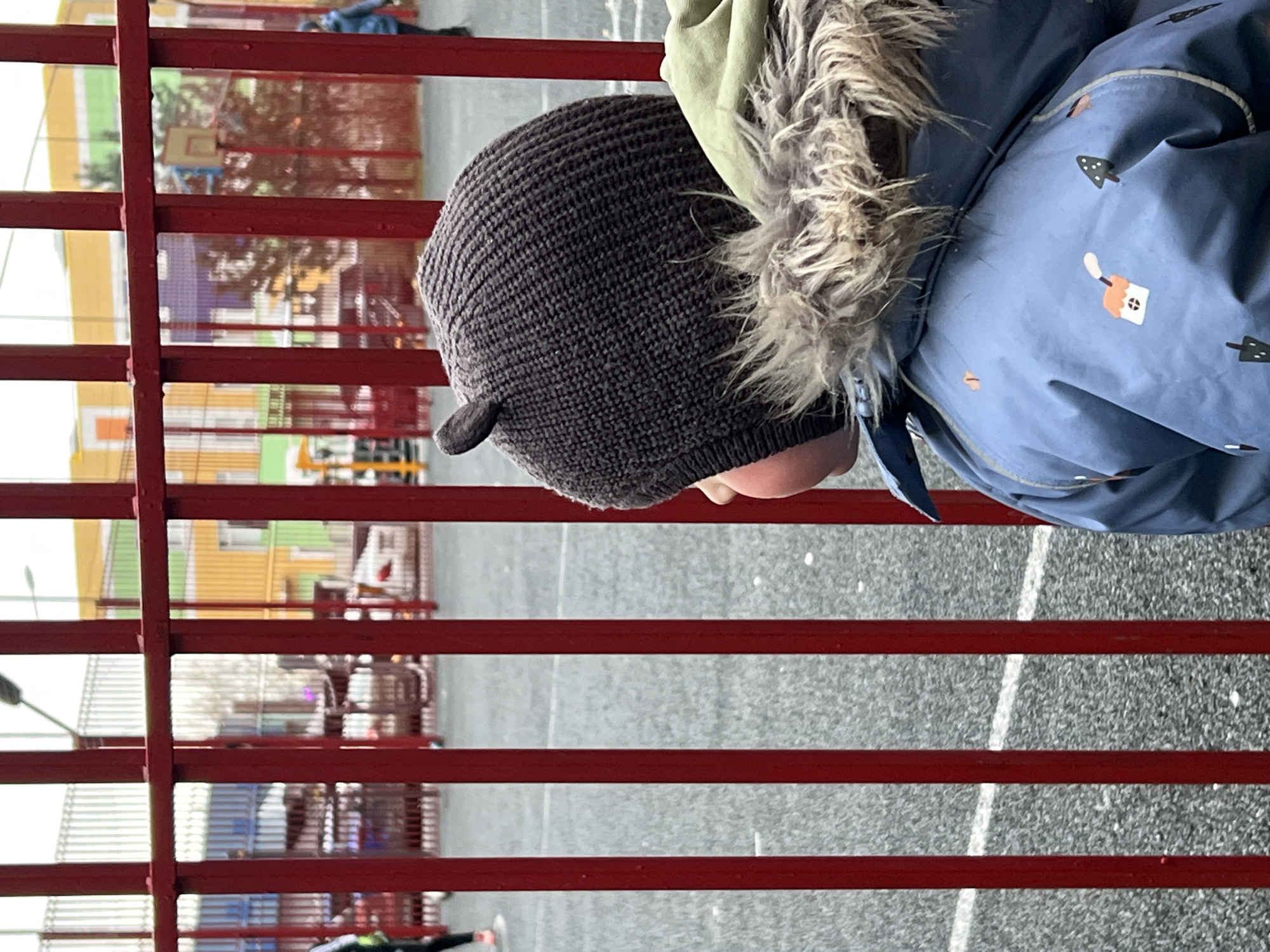 A child is standing near a red metal fence. The child is wearing a knitted hat with ear-like protrusions, a fur-lined jacket with a hood, and a blue jacket with cartoon-like designs. Through the fence, a playground area with a basketball hoop and a building with yellow, green, and red colors can be seen. The ground is grey. Based on the buildings and playgrounds visible through the fence, the image was taken in the city of Khimki, Russia.