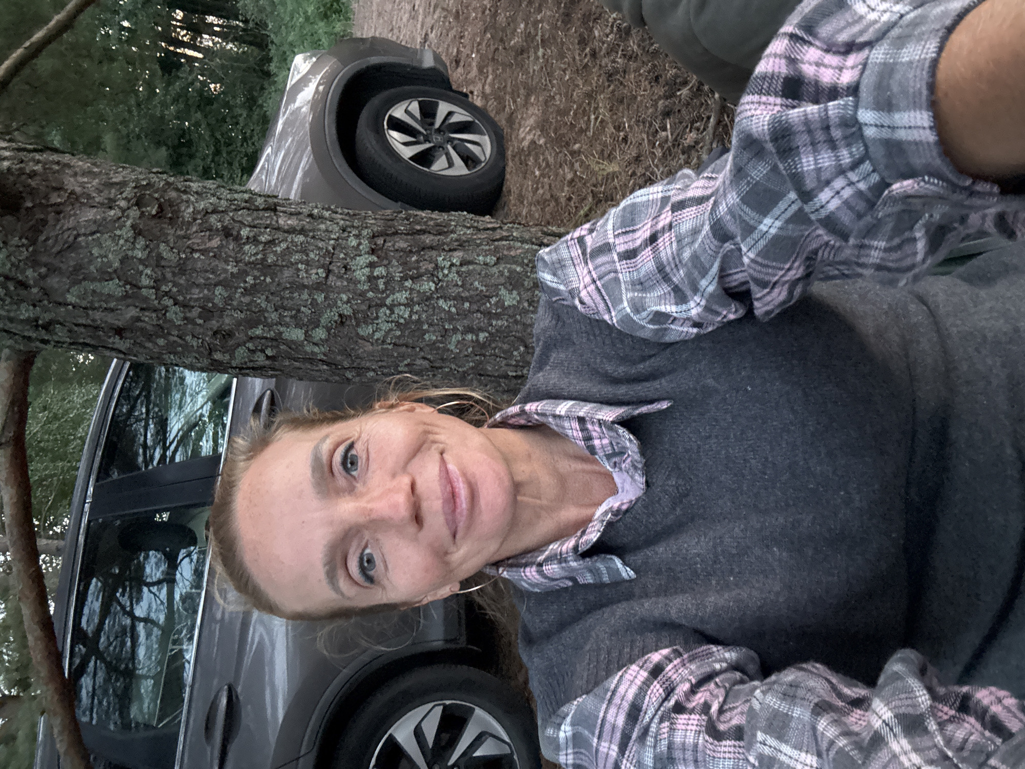 A woman is taking a selfie outdoors. She is sitting in front of a tree, with a car parked behind her. The woman has blonde hair and blue eyes and is wearing a gray sweater over a plaid shirt. The car is a dark color and appears to be an SUV. The environment is wooded with some undergrowth. It is daytime, and the lighting suggests it may be late afternoon or early evening. There are no visible signs or landmarks to indicate the specific location in Zelenaya Roshcha, Russia, beyond it being an outdoor, possibly park-like or forested area.