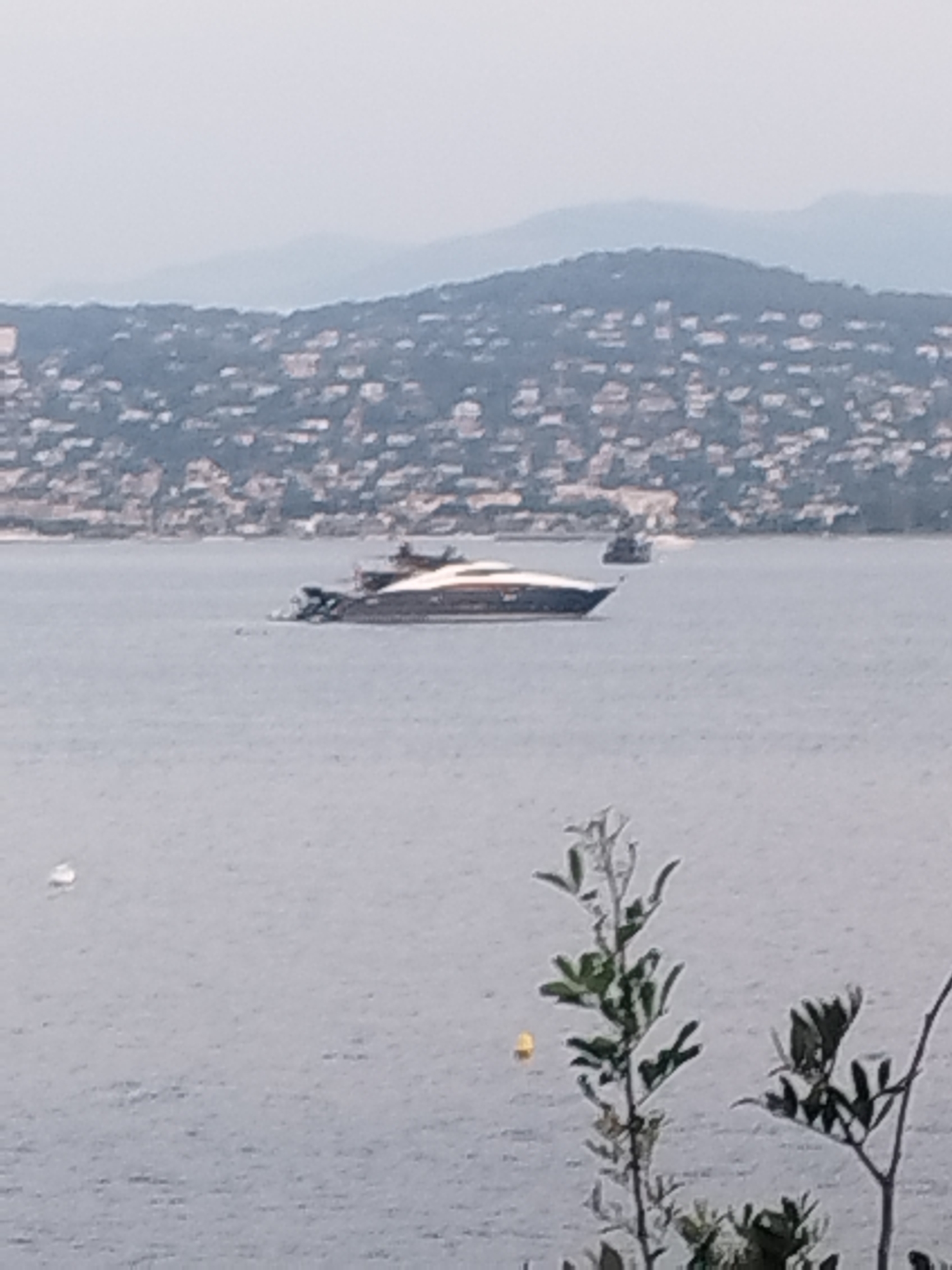 A panoramic view captures a distant, sprawling coastline of Antibes, France, dotted with buildings and hills. The foreground is dominated by a large, sleek yacht navigating the calm, grey waters of the sea. In the background, a smaller boat is visible, adding to the maritime scene. The sky is overcast, contributing to the muted color palette of the image.