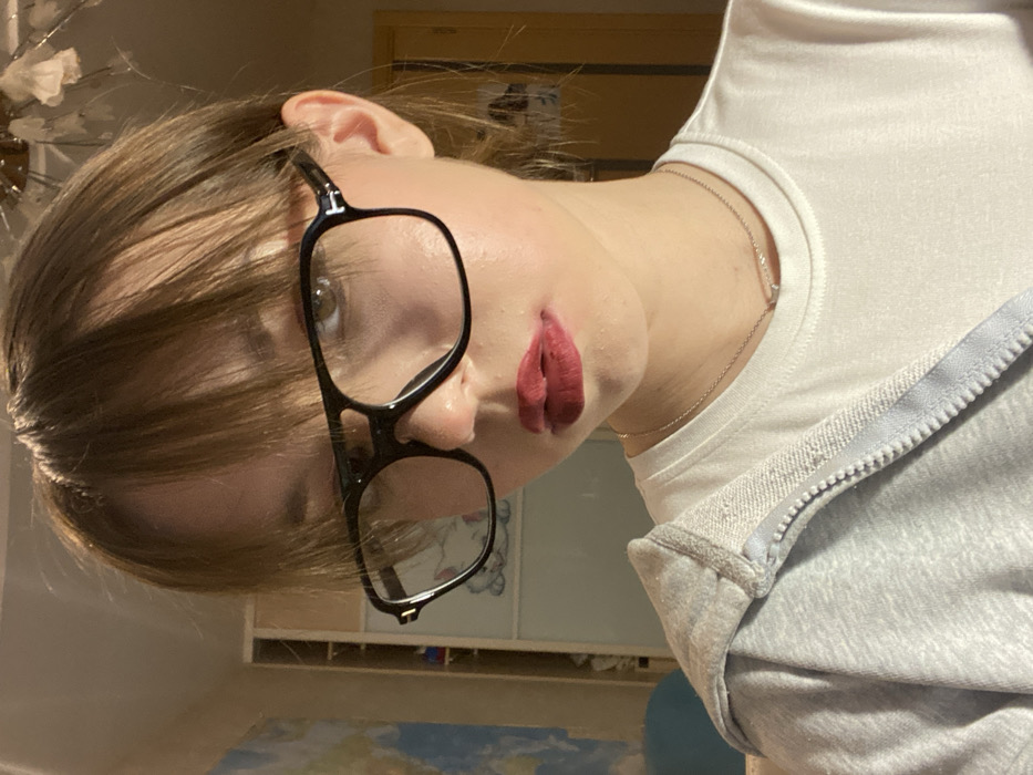 The image captures a waist-up, slightly upward-angled selfie of a young person in an indoor residential setting in the city of Chaikovsky, Russia. The subject, appearing to be female with fair skin, has medium brown hair styled with bangs covering the forehead. She is looking directly into the camera with brown eyes, wearing large black-framed eyeglasses featuring a subtle 'T' logo on the temples. Her lips are coated with dark red lipstick. She wears a plain white short-sleeved t-shirt and a light grey zippered hoodie, open and draped over her left shoulder. A thin silver necklace with a small, horizontal bar pendant is visible around her neck.
The background reveals elements of a room. To the left, a light-colored wall features a large, partially visible graphic or map, and a portion of a light blue spherical object. Further left, a vertical piece of light-toned wooden furniture is discernible. Towards the right, a white wardrobe or cabinet with two paneled doors is visible, one door displaying a partially visible outline of a white cat illustration. Above, a decorative ceiling light fixture, possibly a chandelier with translucent, flower-like elements, is present. A vertical strip of yellow-colored wall or doorframe is also visible on the far right. The room appears to be illuminated by indoor lighting.